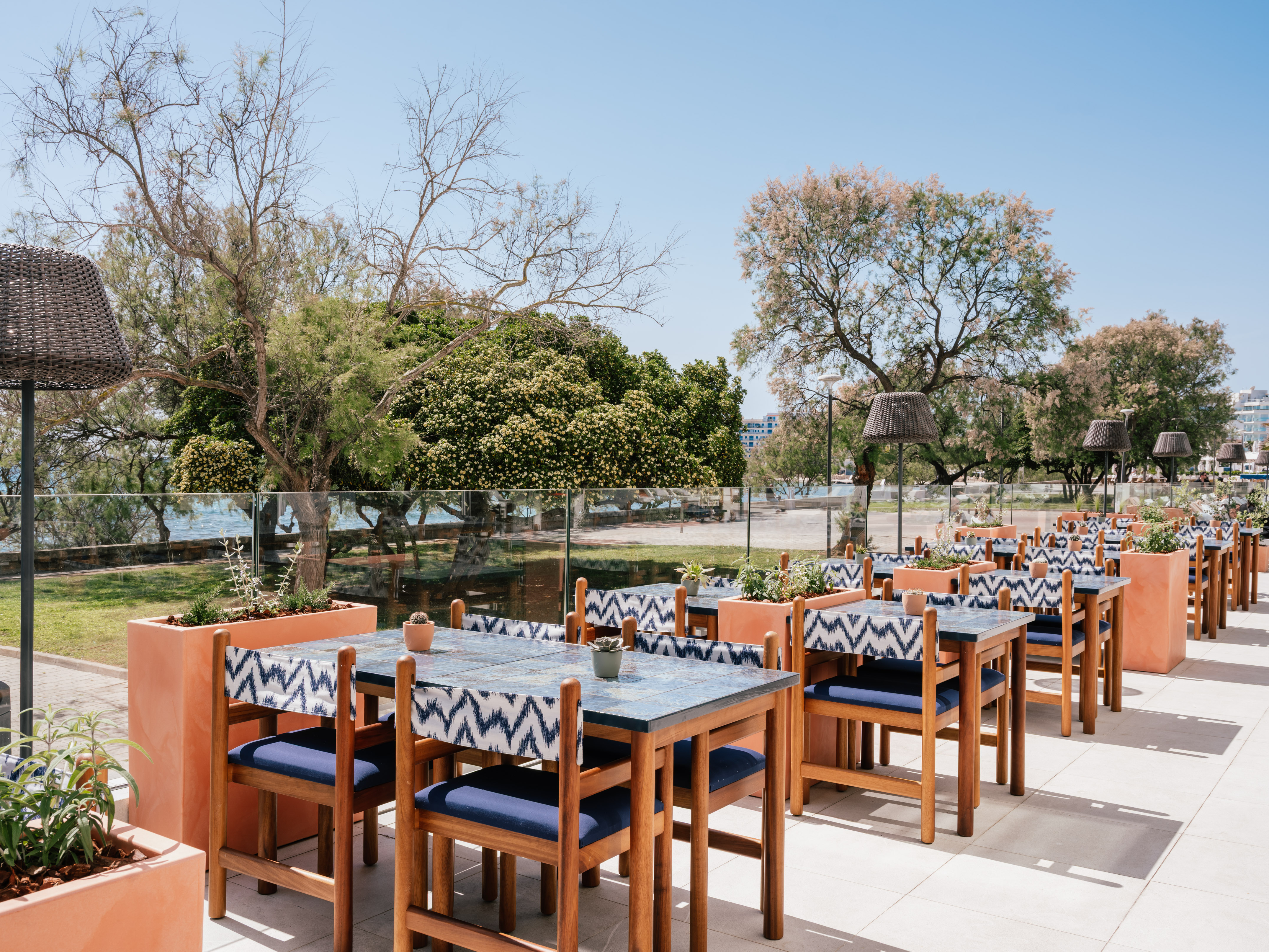 Sunny outdoor restaurant patio with blue tables, patterned chairs, sea view.