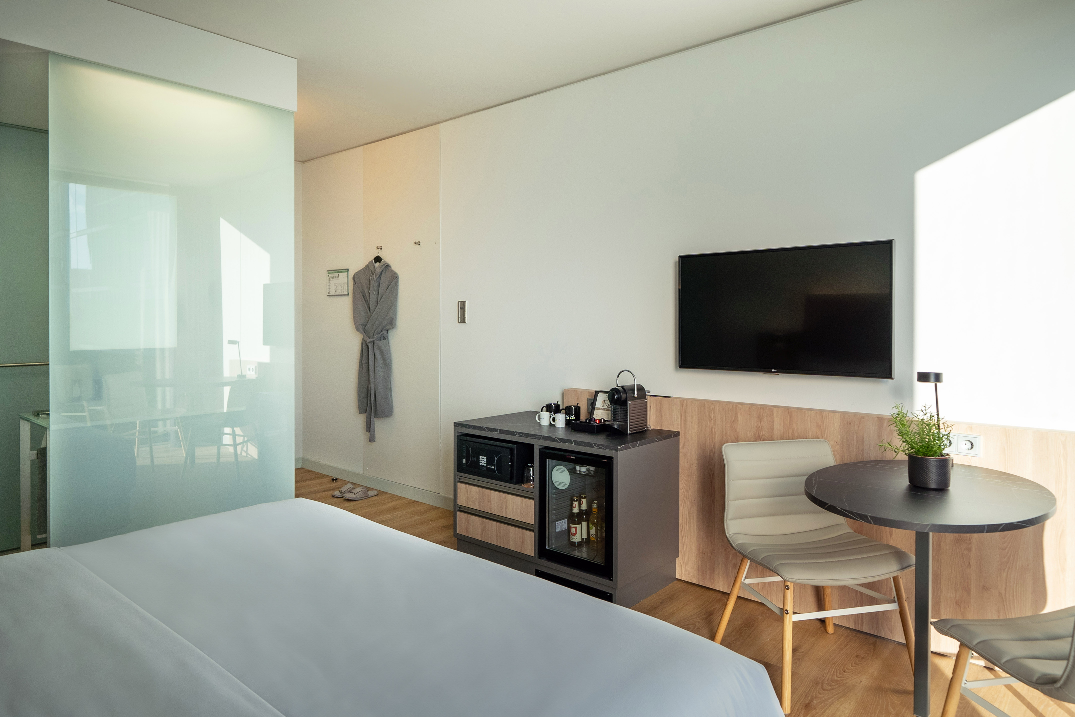 Modern hotel room with white bed, flat-screen TV, minibar, and glass wall.