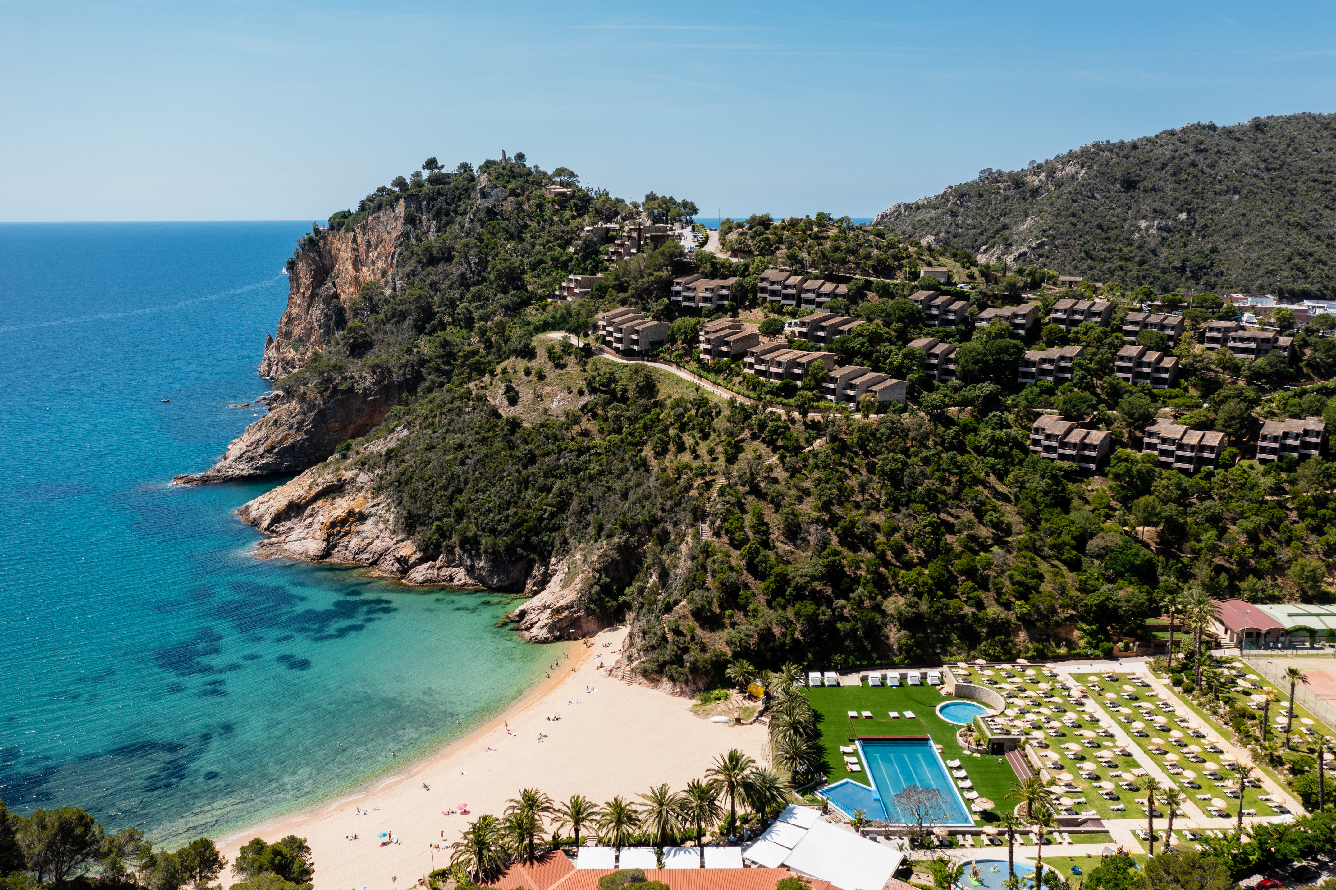 Scenic coastal resort with beach, turquoise sea, hillside villas, and swimming pools.