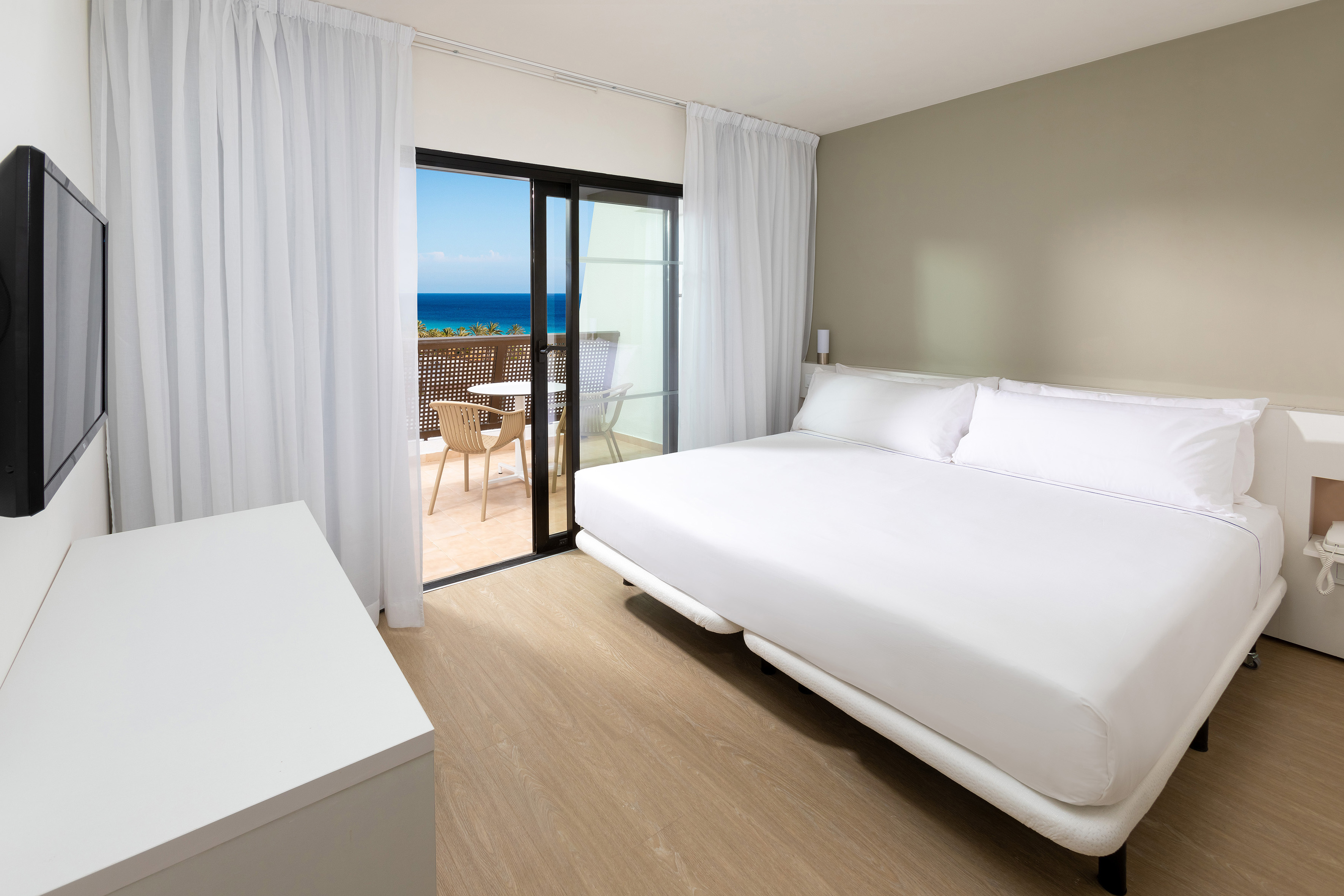 Modern hotel room with ocean view balcony and white bed.