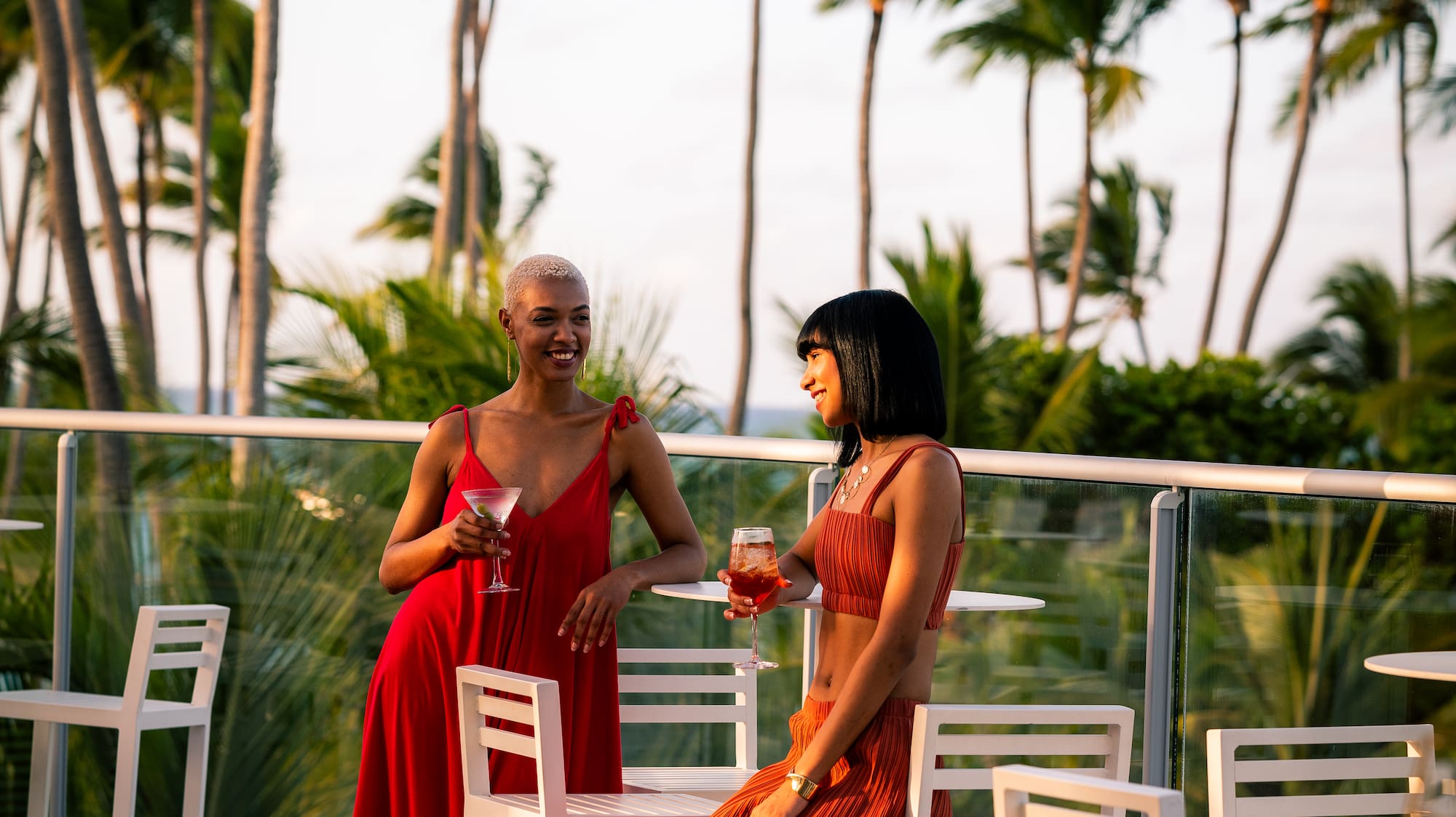 Stylish women enjoying cocktails on a palm-lined resort balcony.