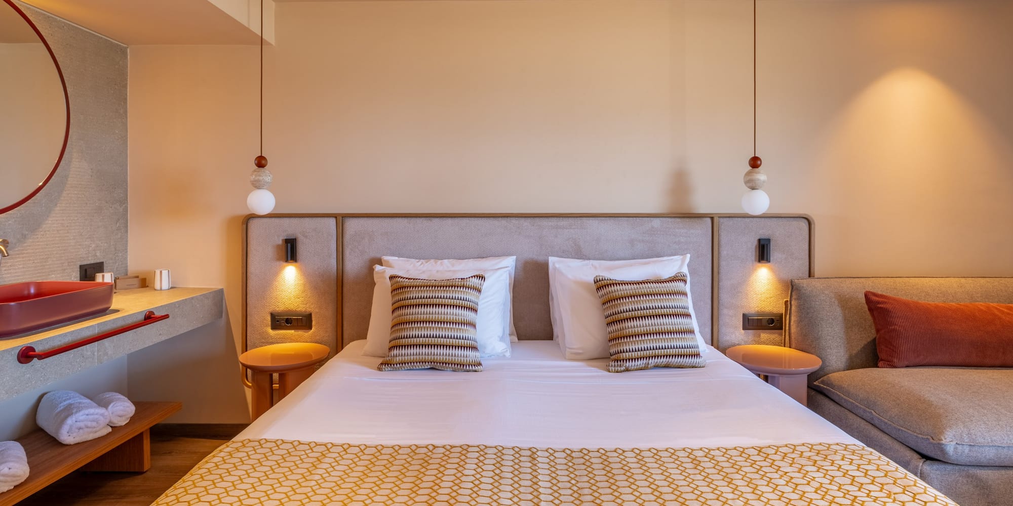 Modern hotel bedroom with large bed, geometric throw, red sink, and lounge.