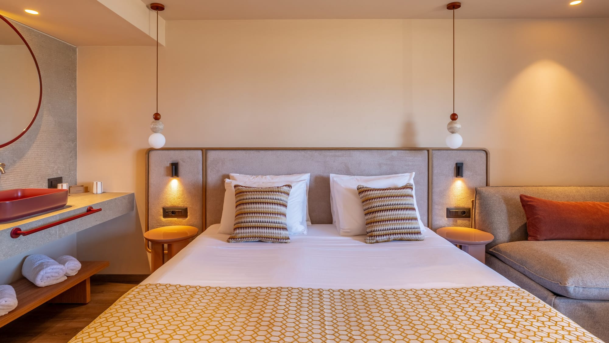 Modern hotel bedroom with large bed, geometric throw, red sink, and lounge.