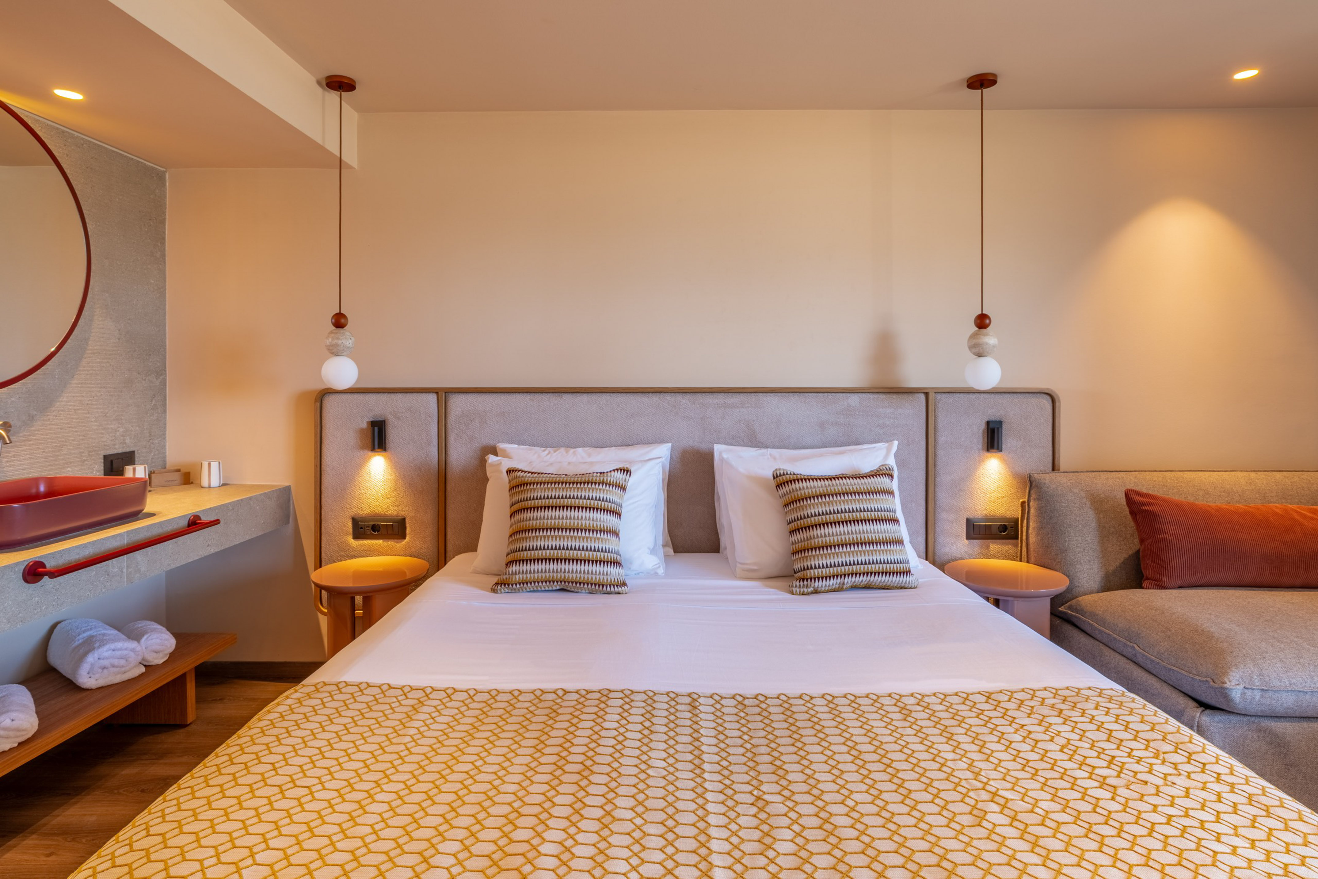 Modern hotel bedroom with large bed, geometric throw, red sink, and lounge.
