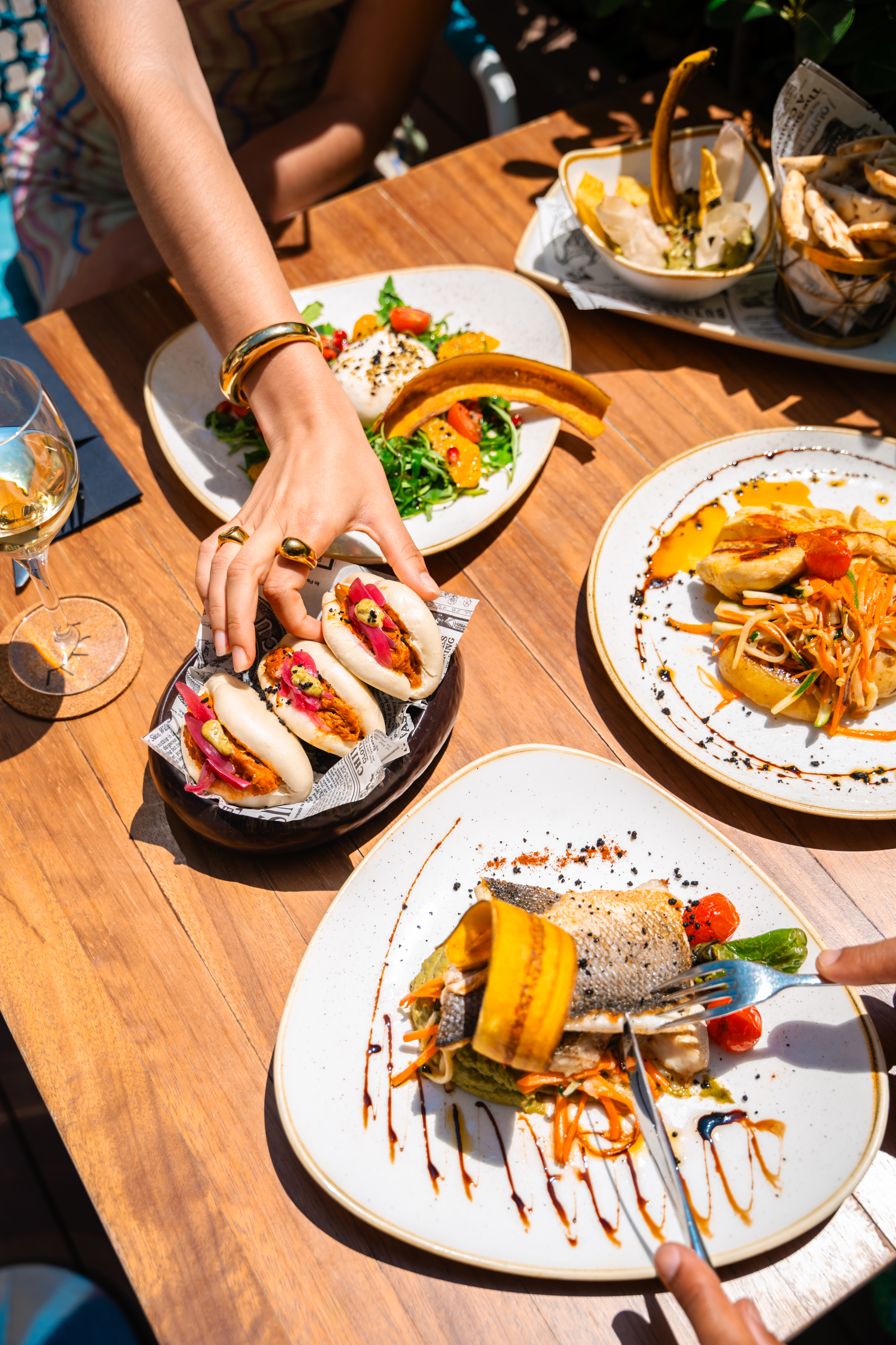 Gourmet dining with bao buns, fish, and burrata salad on a sunny wooden table.