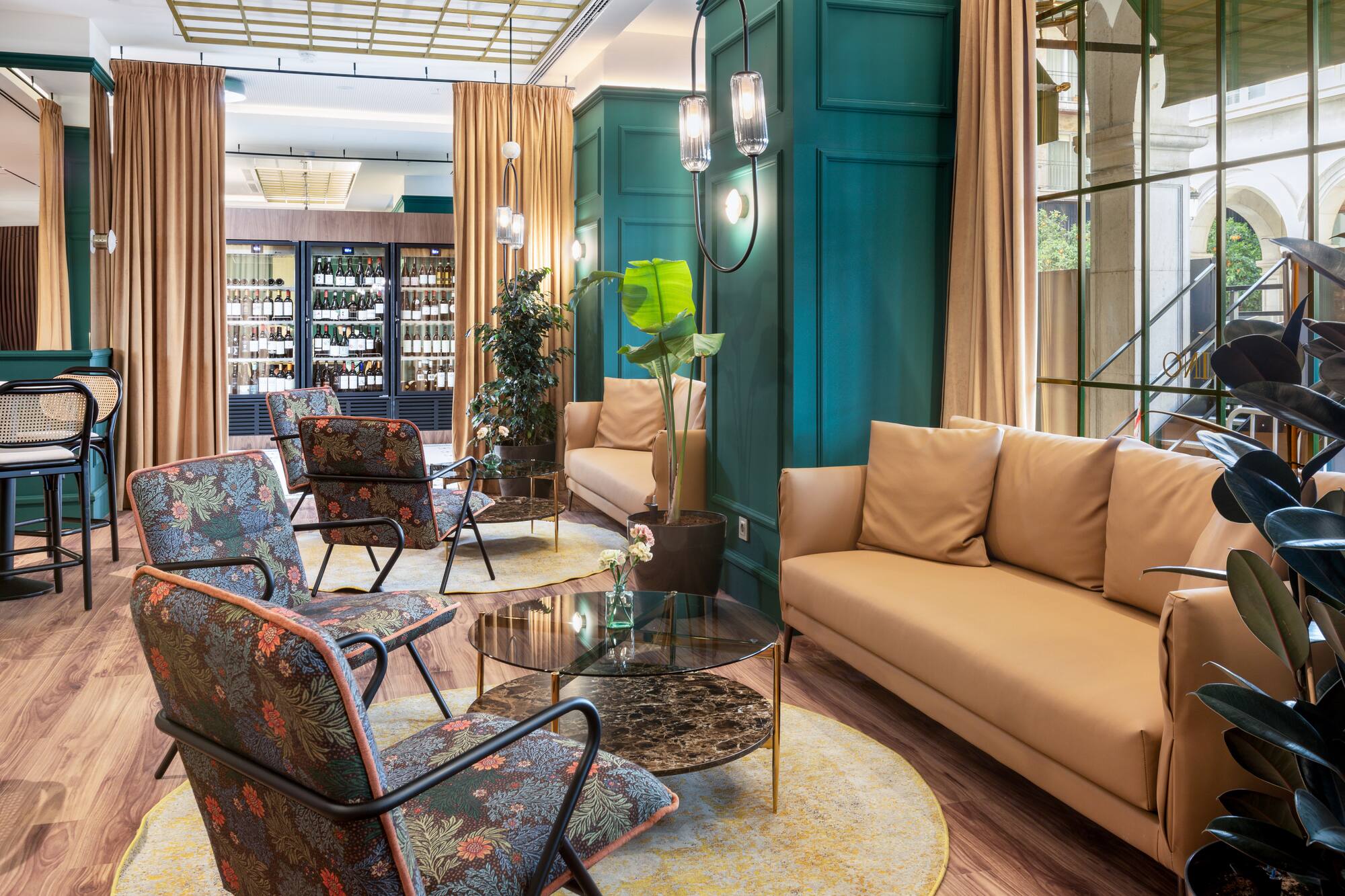 Teal-paneled hotel lounge with tan sofas, floral chairs, and wine display.