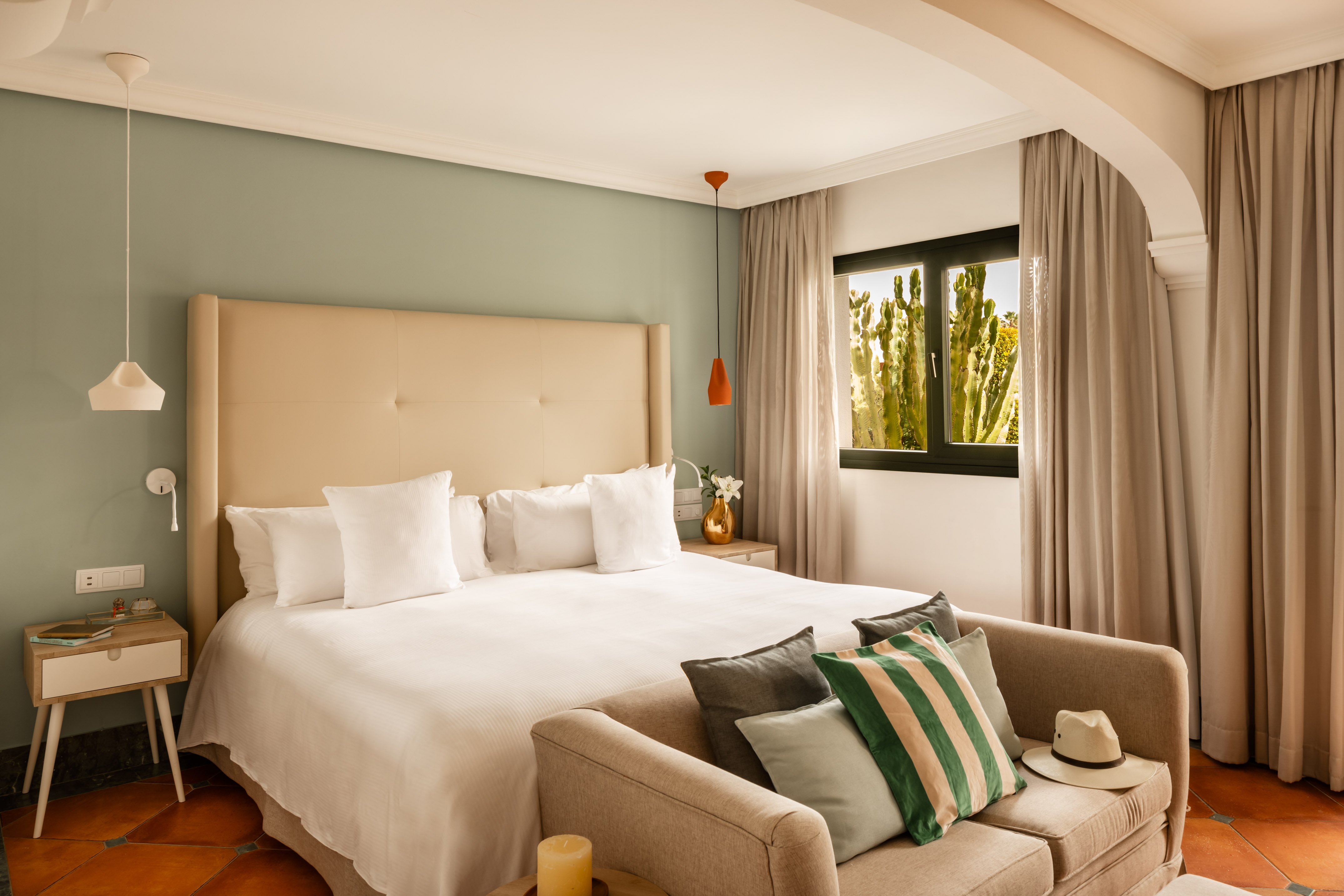 Bright bedroom with large bed, green wall, beige sofa, and window showing cacti.