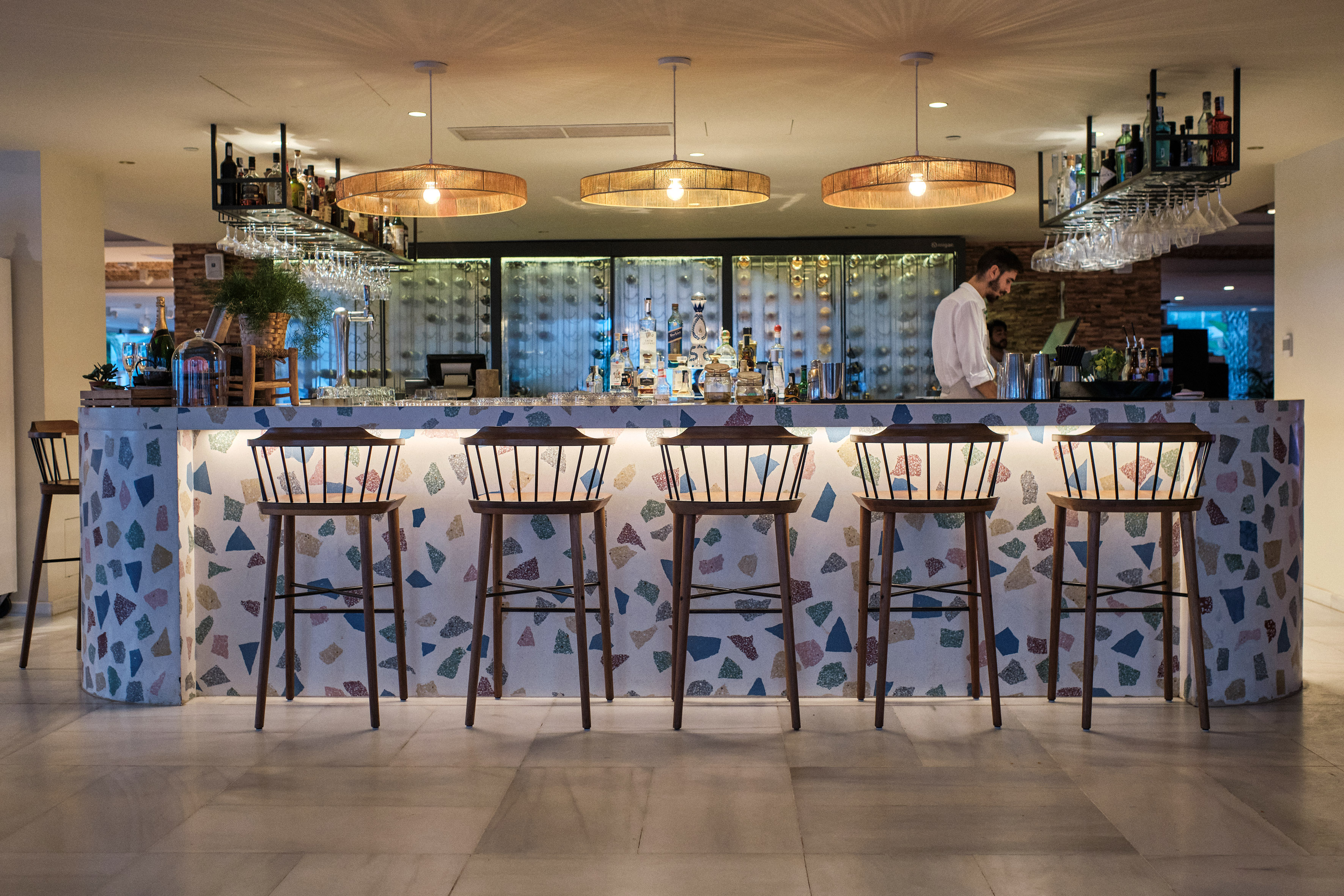 Modern bar with colorful terrazzo counter, wooden stools, and bartender.