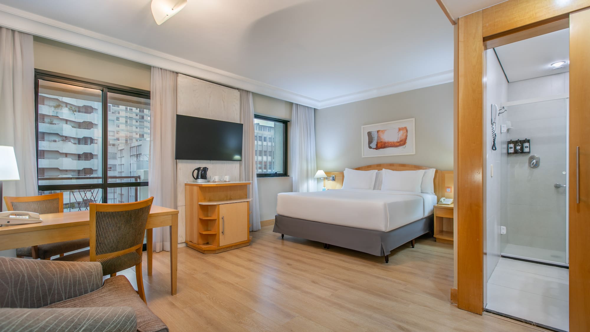 Modern hotel suite with king bed, desk, city view, and shower.