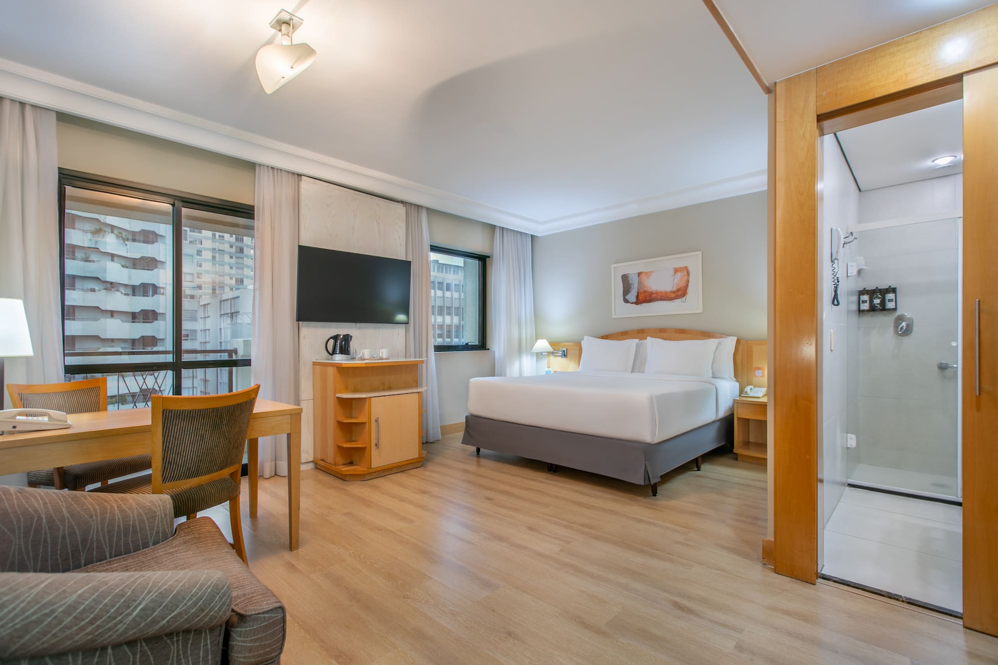 Modern hotel suite with king bed, desk, city view, and shower.
