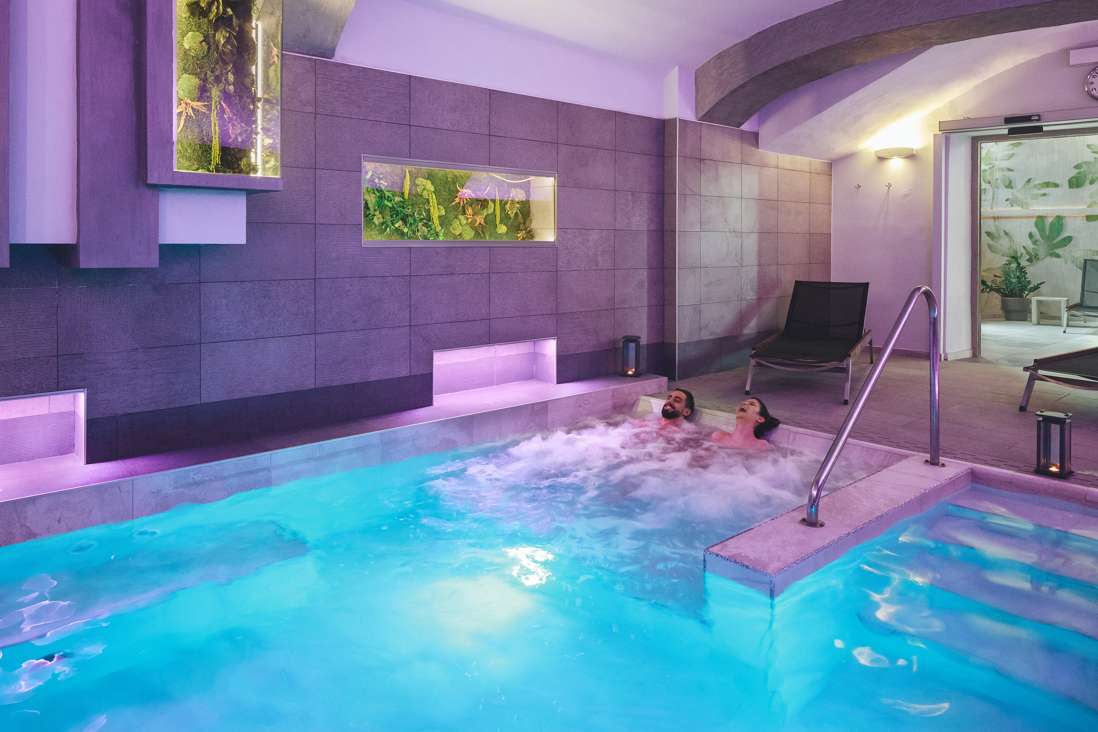 Relaxing couple in a luxury spa jacuzzi with vibrant lighting and plant walls.
