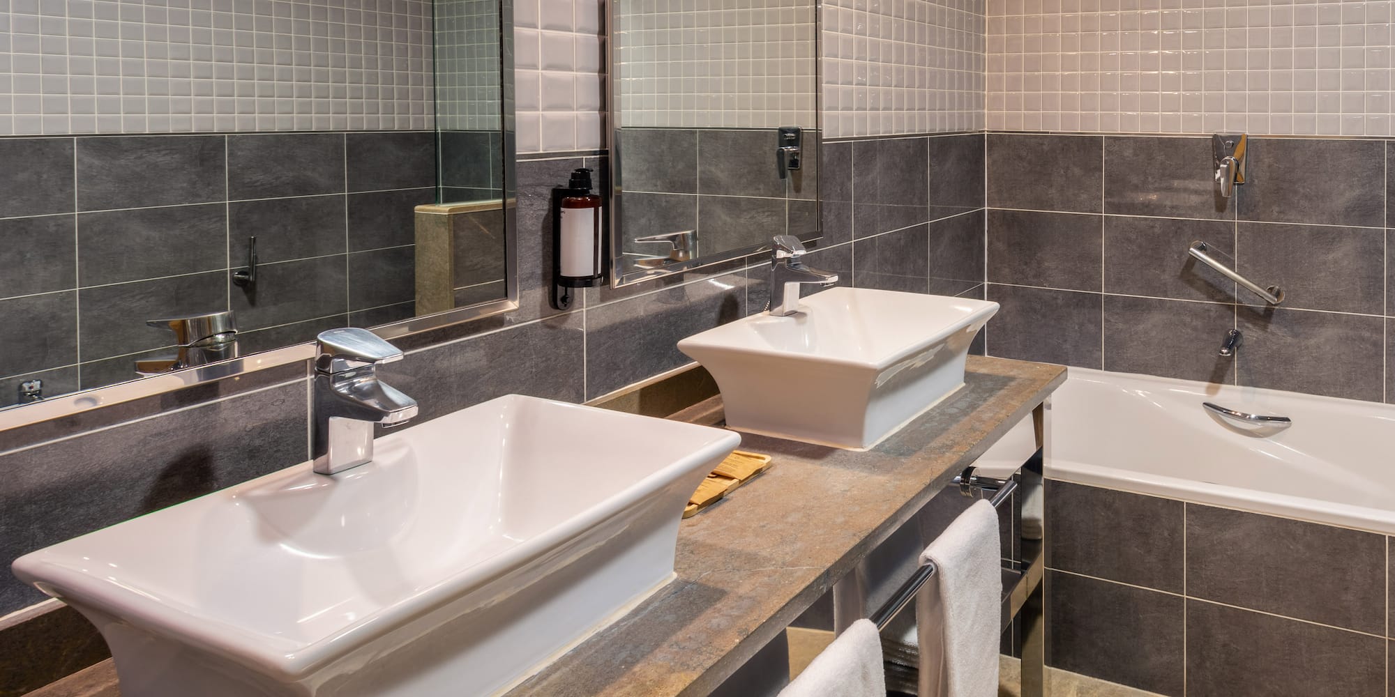Modern bathroom with dual vessel sinks, stone counter, grey tiles, and bathtub.