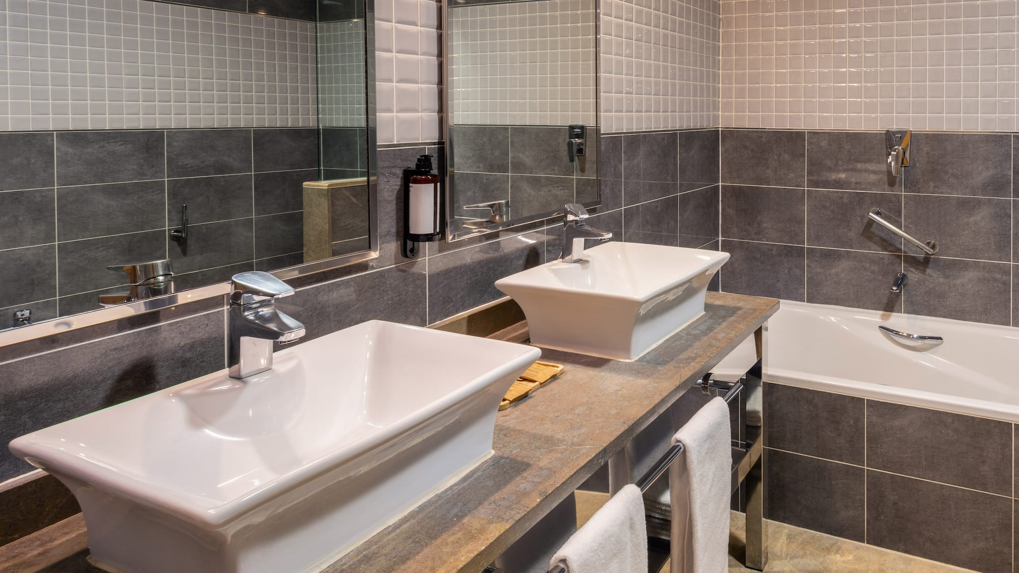 Modern bathroom with dual vessel sinks, stone counter, grey tiles, and bathtub.