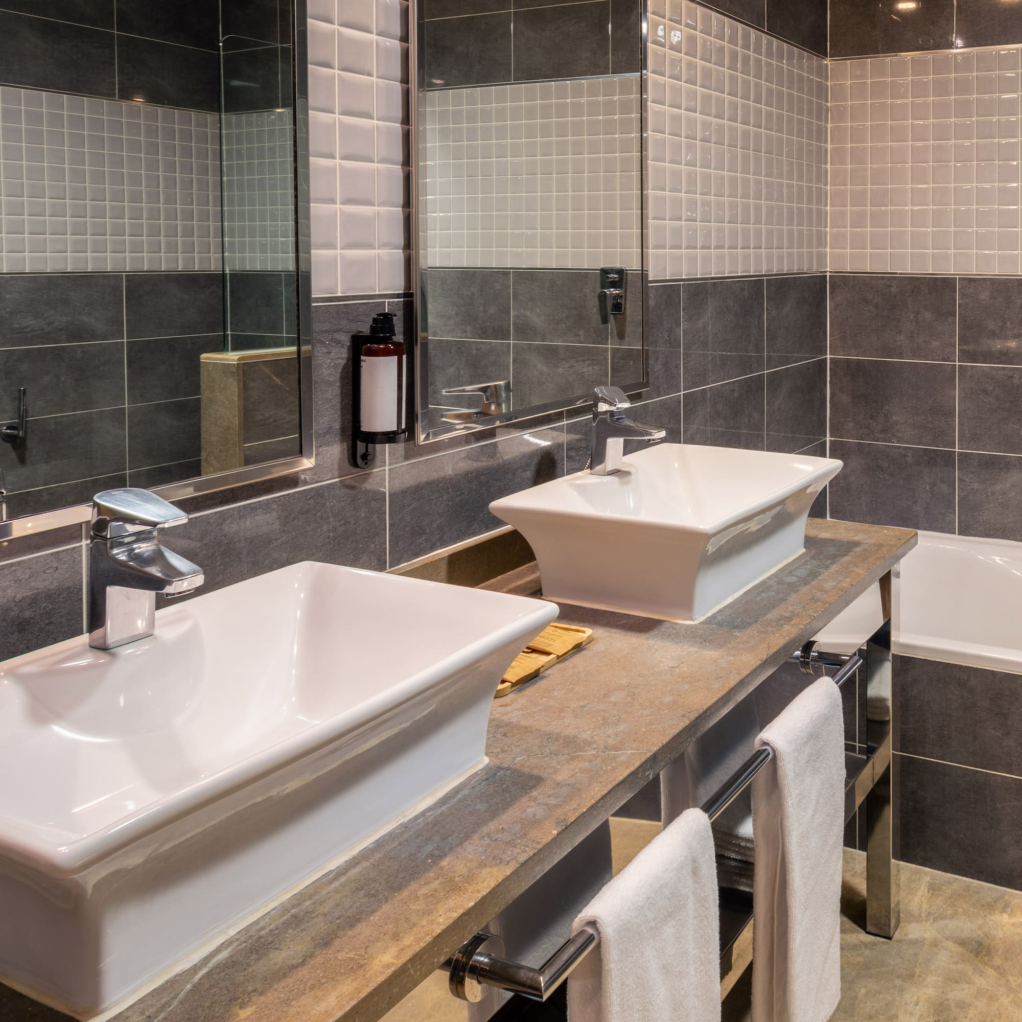 Modern bathroom with dual vessel sinks, stone counter, grey tiles, and bathtub.