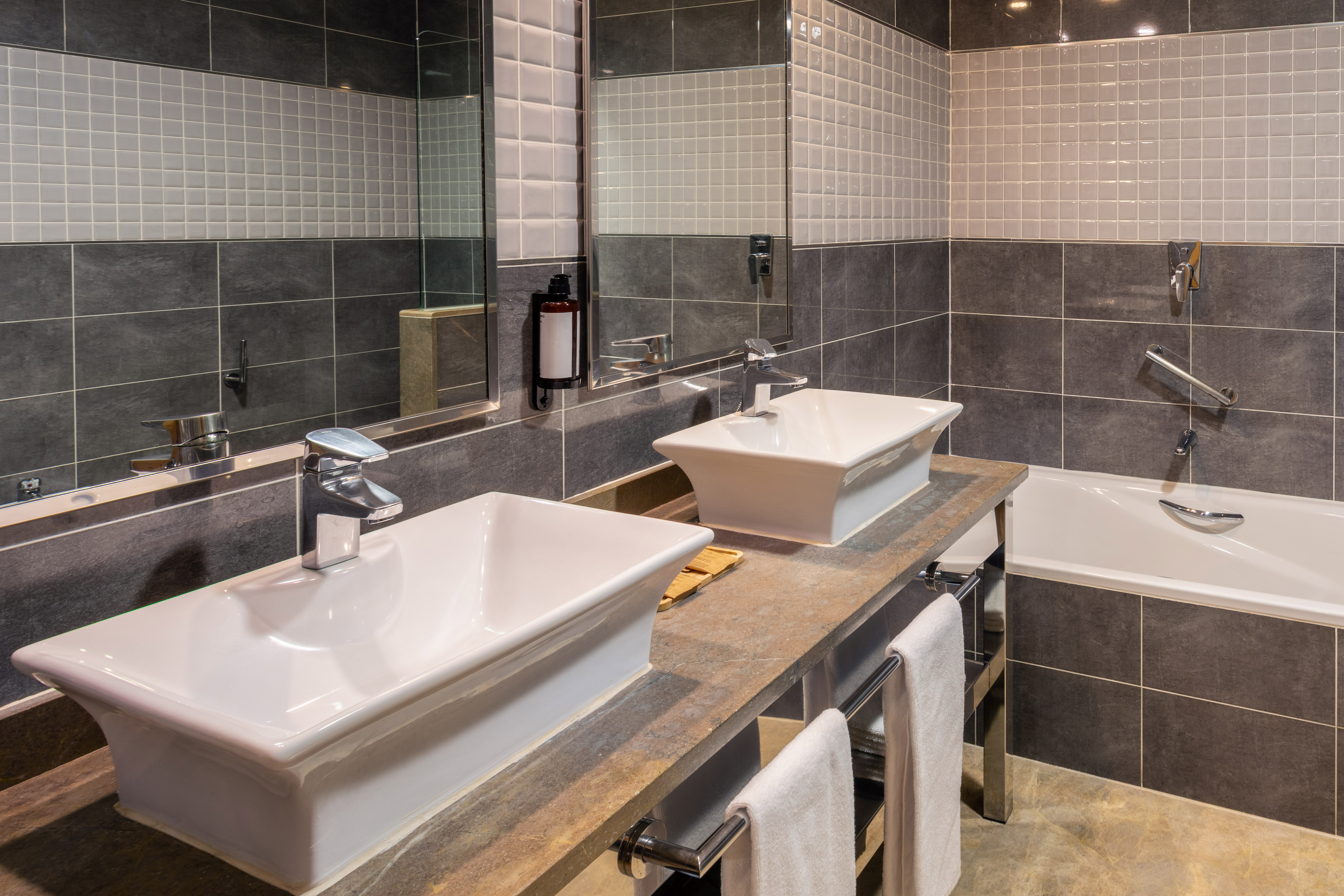 Modern bathroom with dual vessel sinks, stone counter, grey tiles, and bathtub.