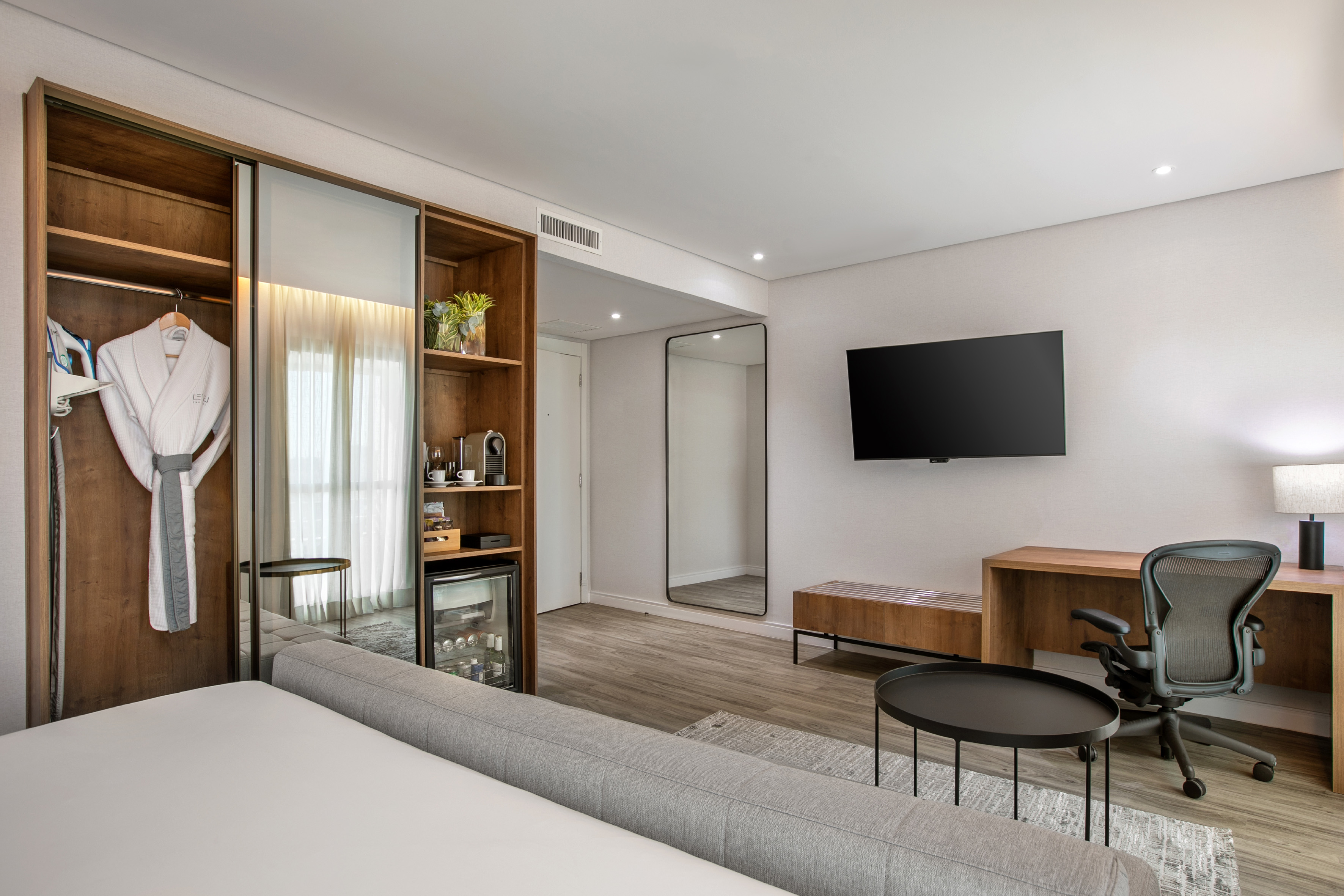 Modern hotel room with large bed, built-in wardrobe, desk, TV, and minibar.