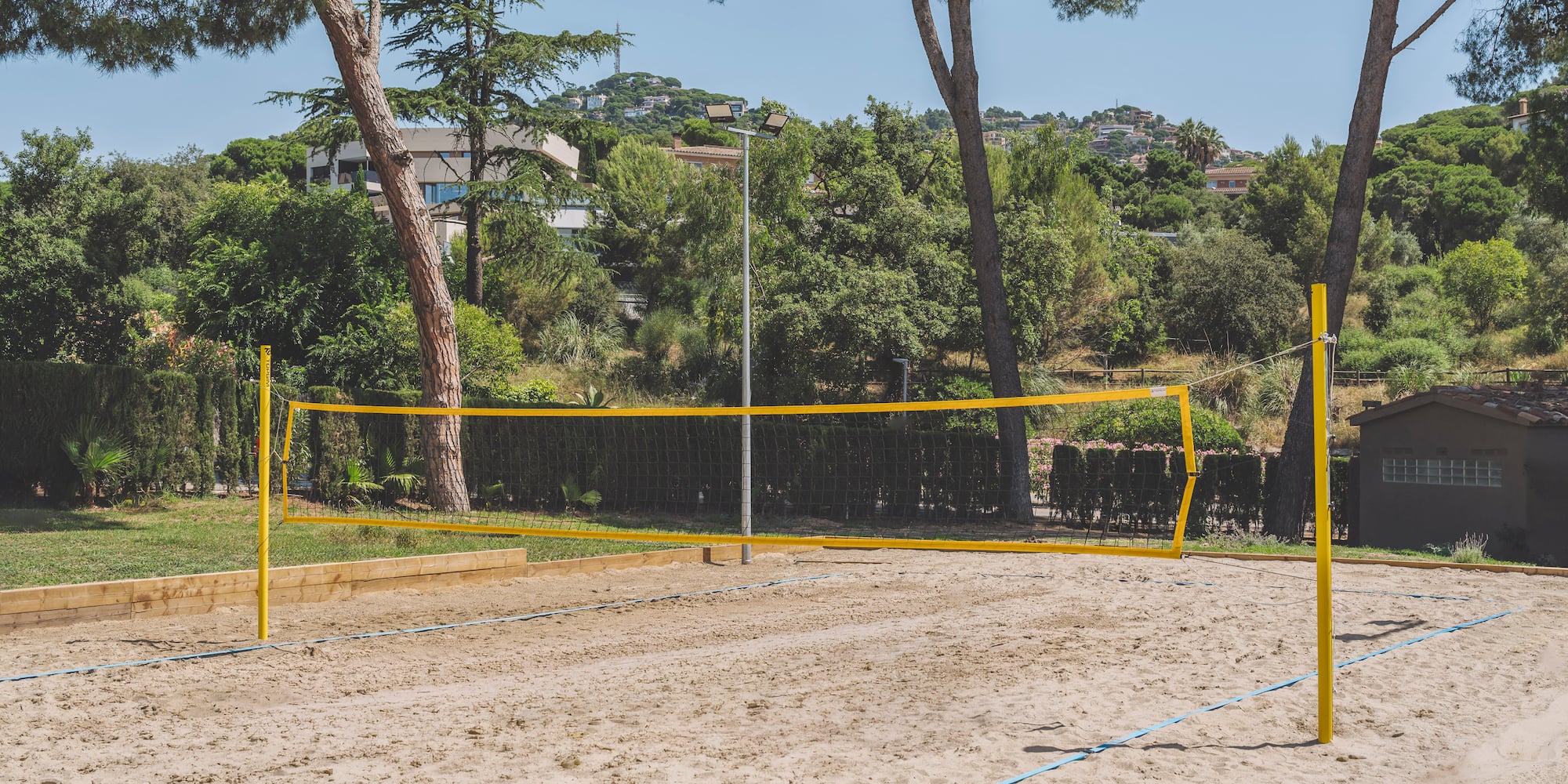 Empty outdoor beach volleyball court on sand with yellow net, surrounded by trees.