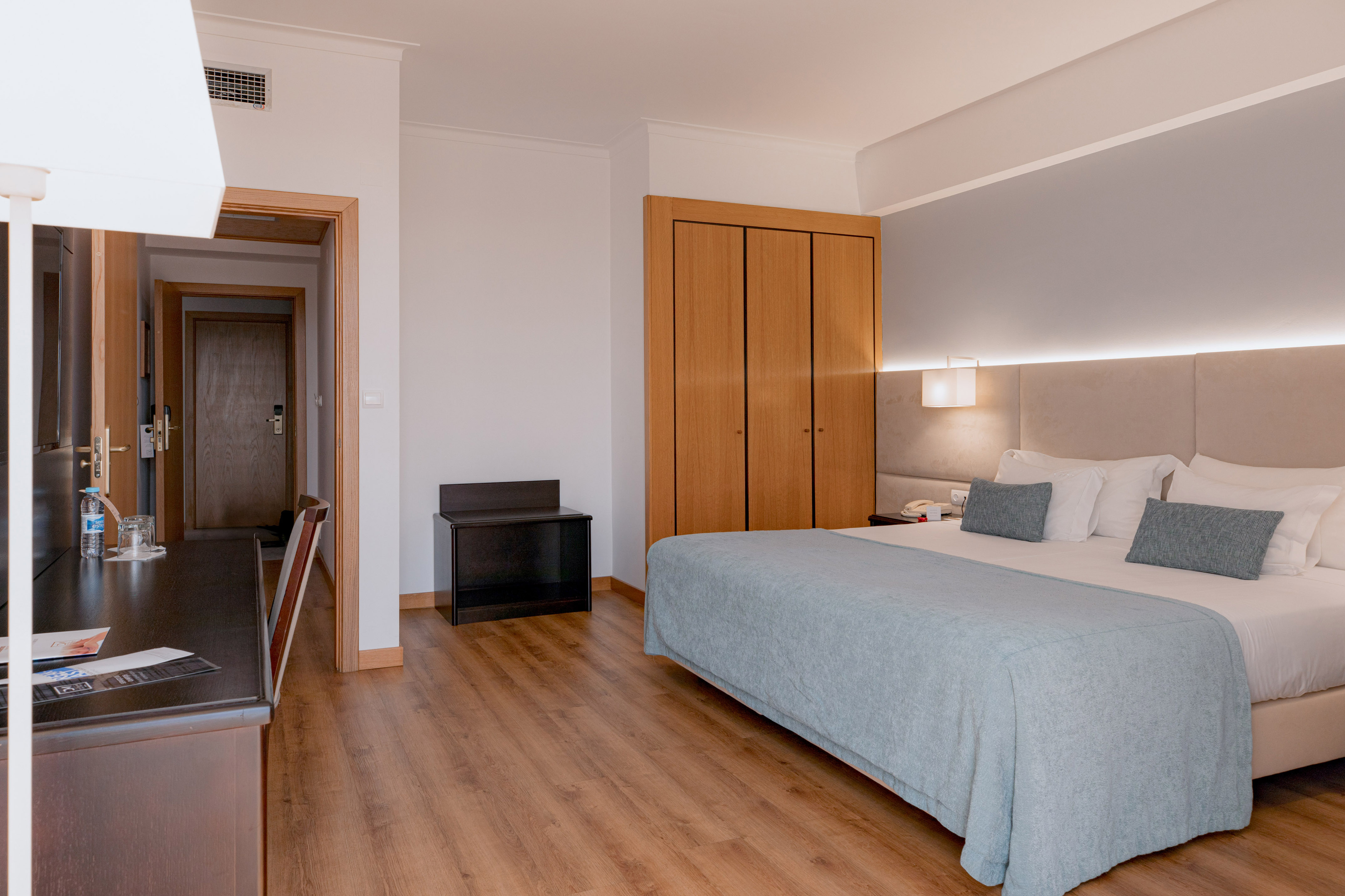 Hotel room: large bed with blue blanket, wood floor, wardrobe, and desk.