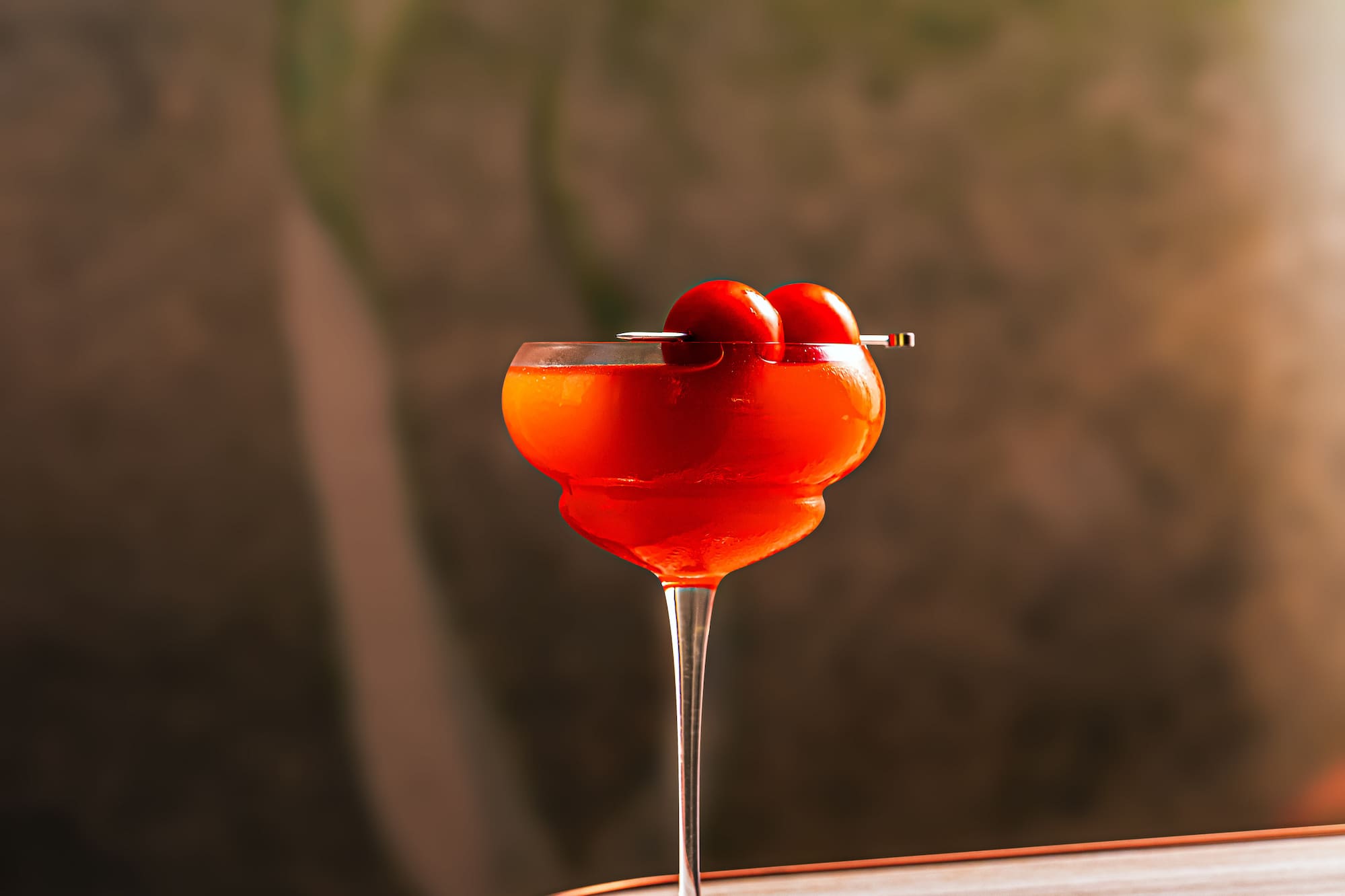 Cóctel rojo intenso en copa, con dos cerezas, sobre mesa de madera.