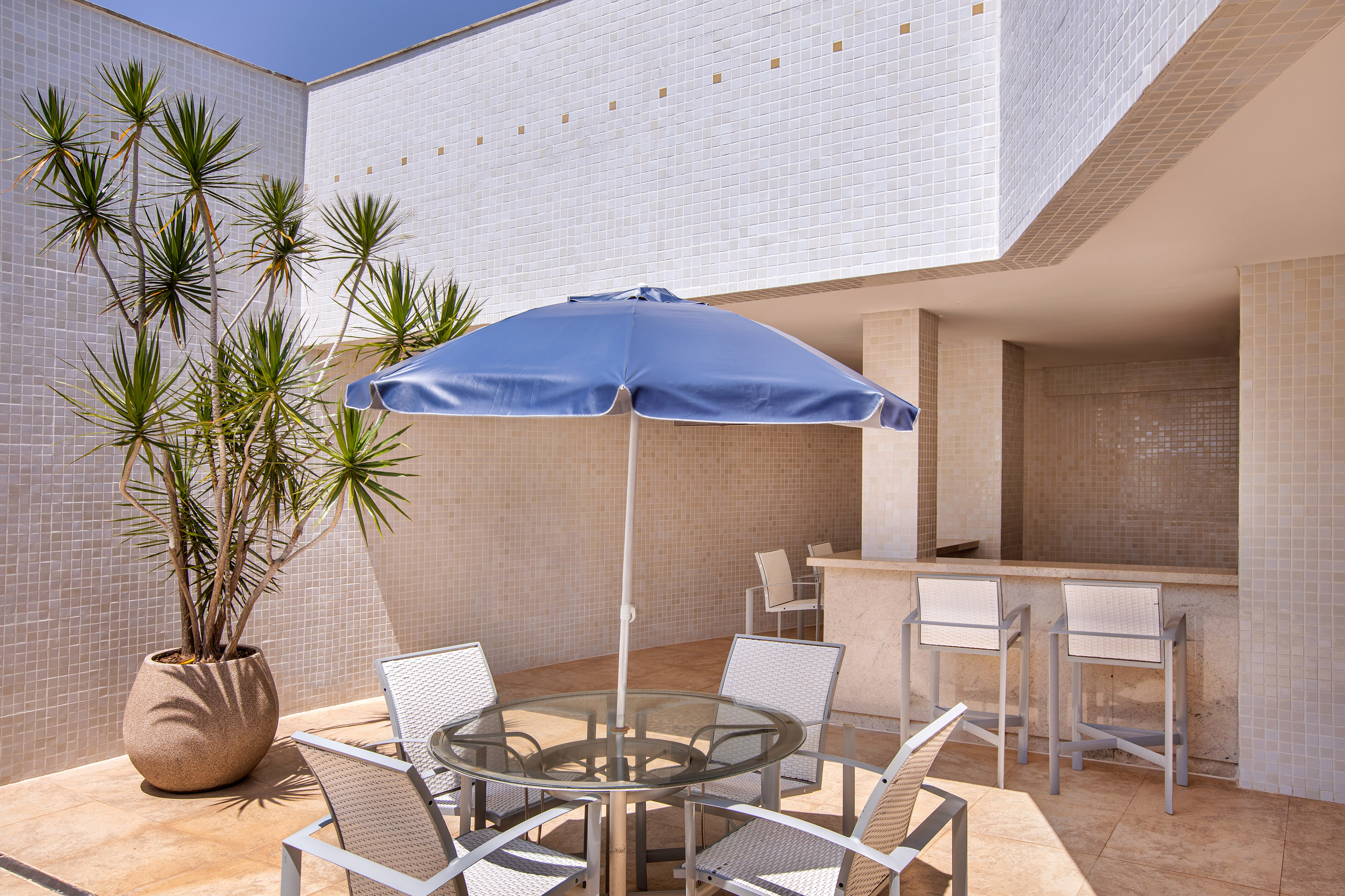 Sunny tiled patio with blue umbrella, outdoor dining set, bar, and potted plant.