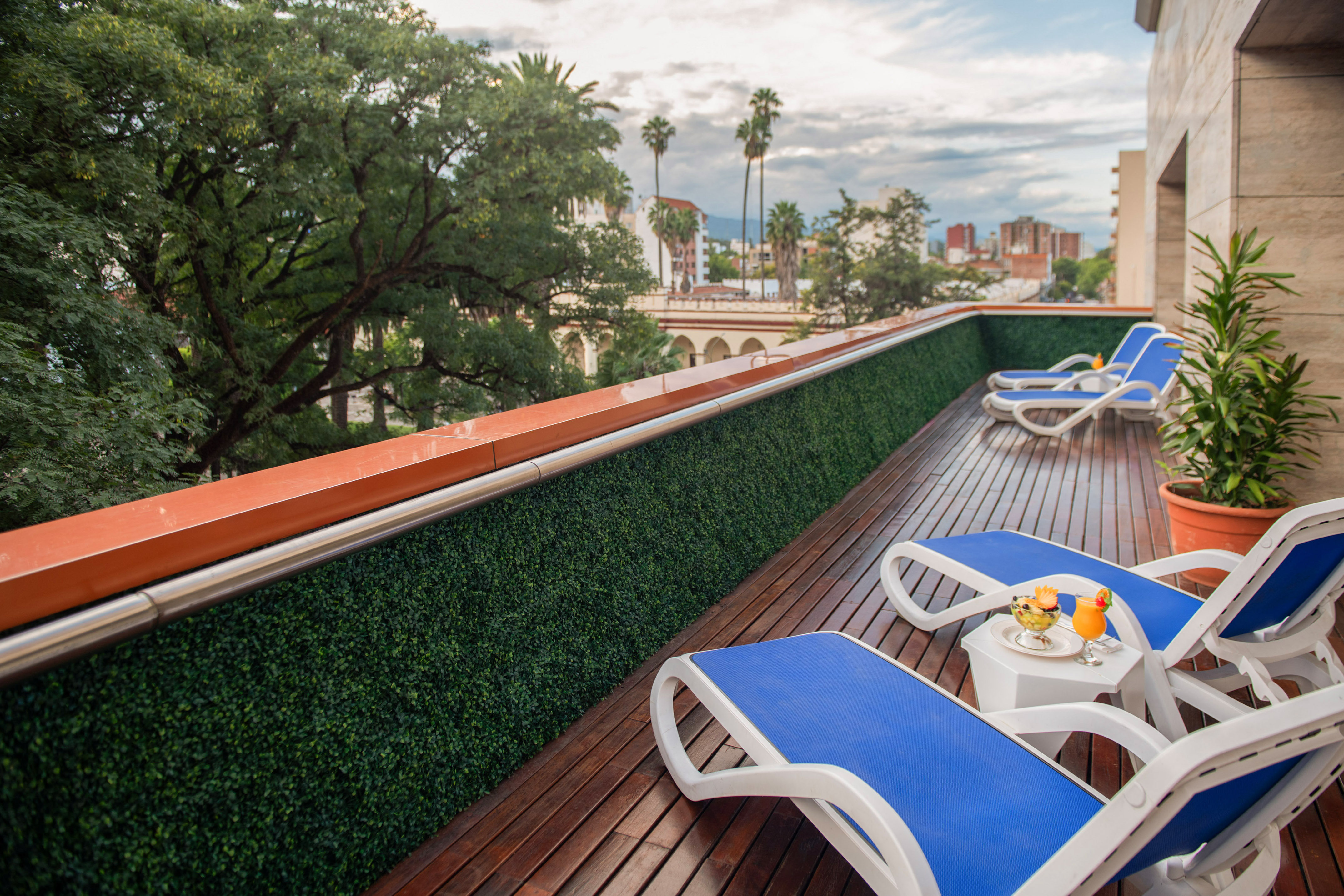 Hotel terrace with blue lounge chairs, wooden deck, green hedge, city view.