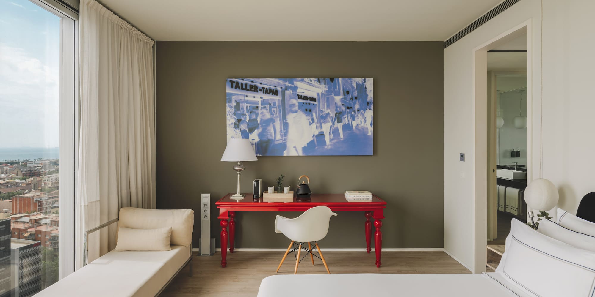 Suite moderna con vista urbana, escritorio rojo, cama y chaise longue.