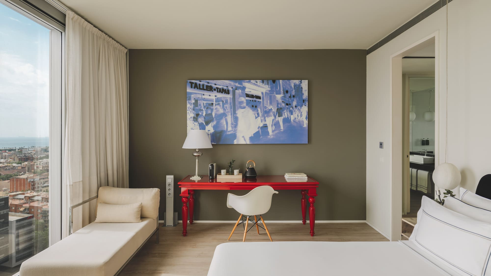 Suite moderna con vista urbana, escritorio rojo, cama y chaise longue.