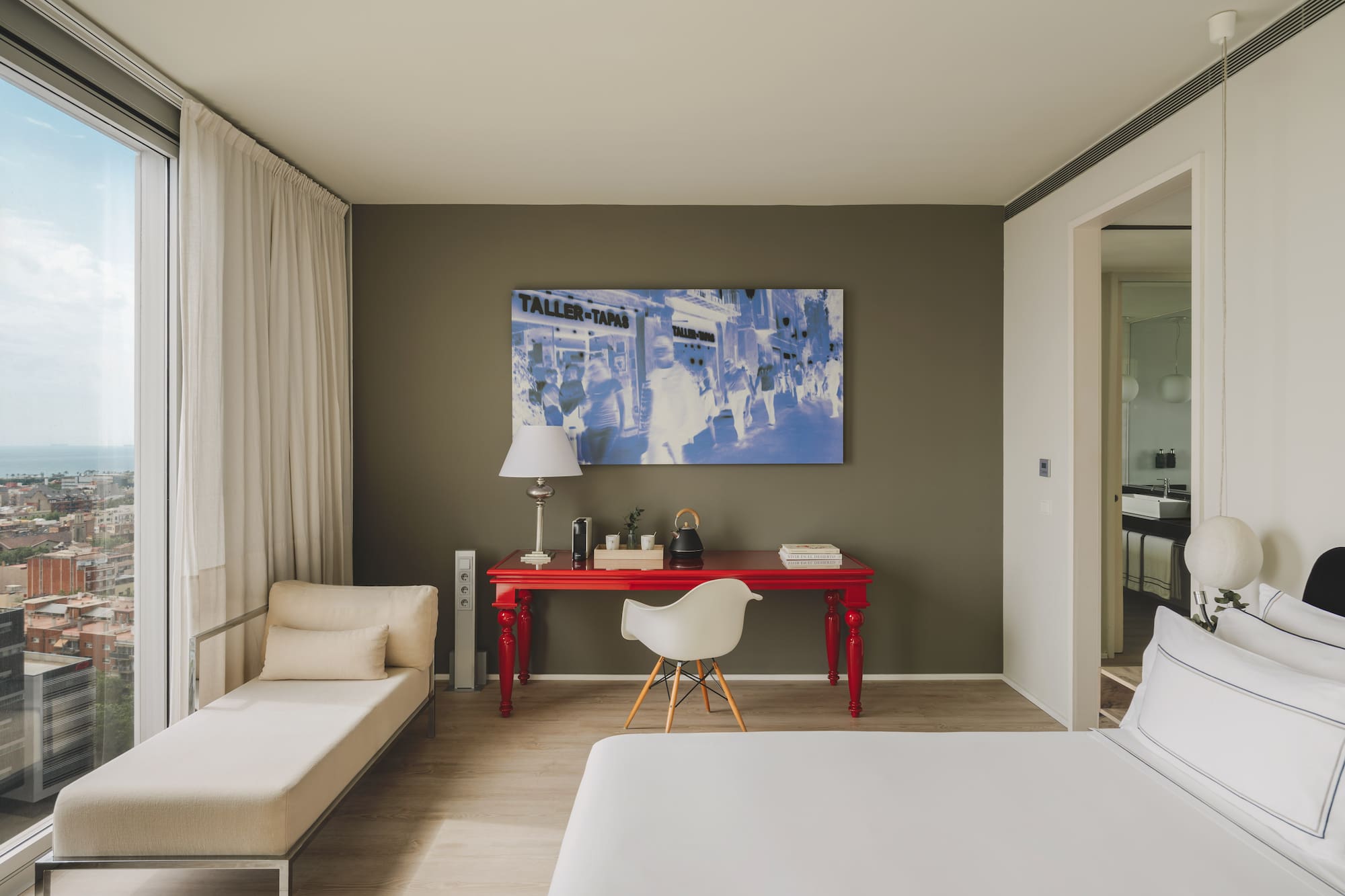 Suite moderna con vista urbana, escritorio rojo, cama y chaise longue.