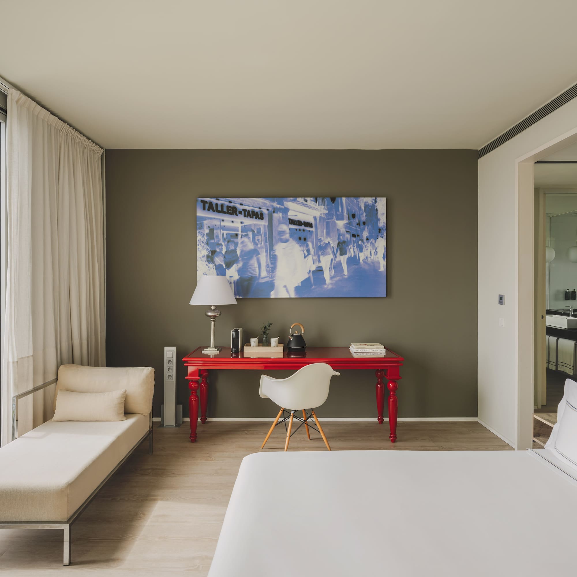 Suite moderna con vista urbana, escritorio rojo, cama y chaise longue.