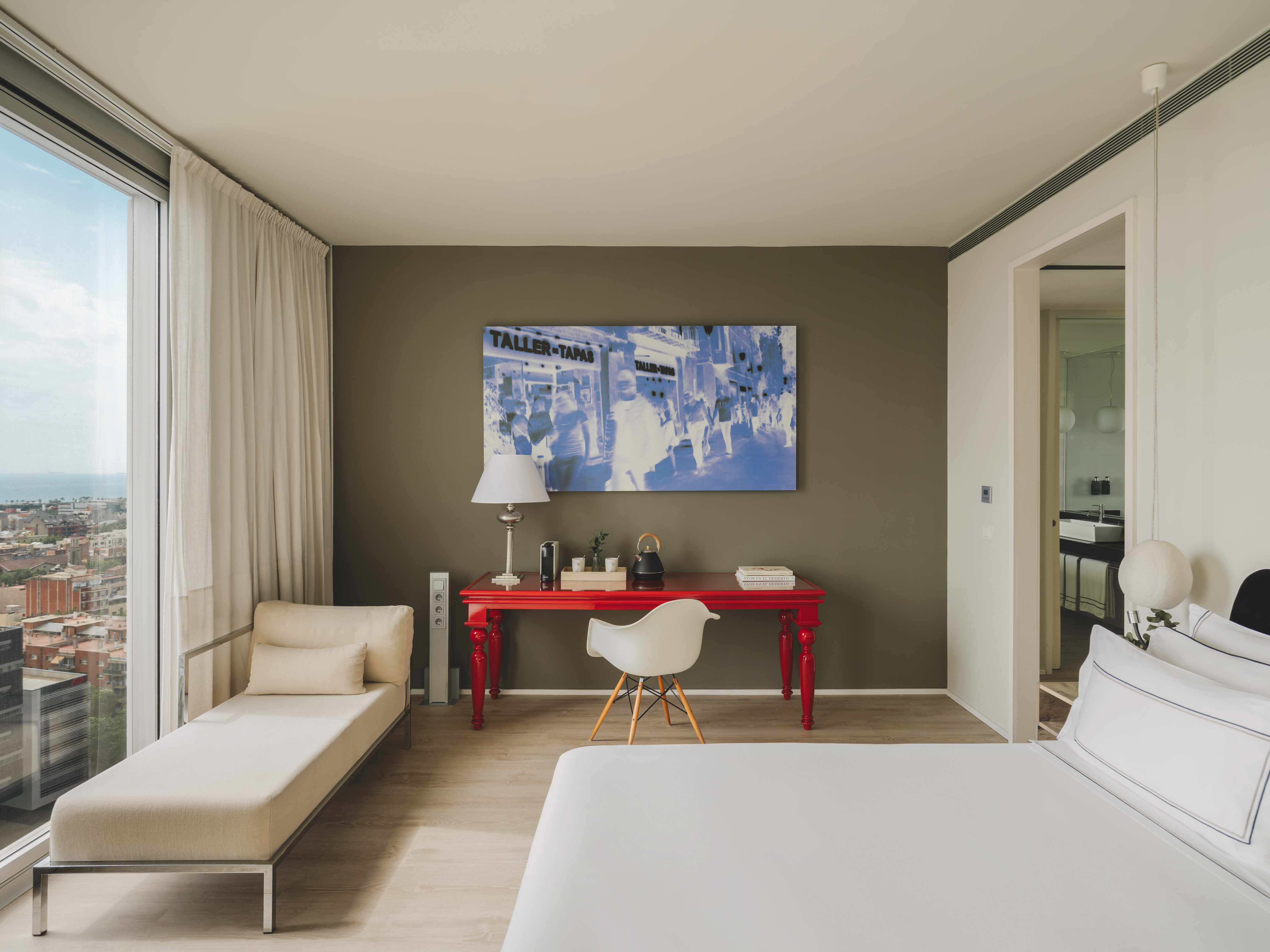 Suite moderna con vista urbana, escritorio rojo, cama y chaise longue.