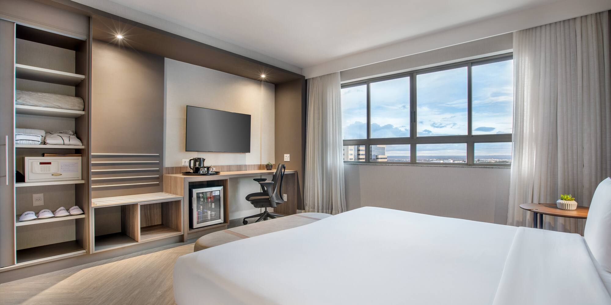 Modern hotel room with bed, workspace, TV, mini-fridge, and city view.