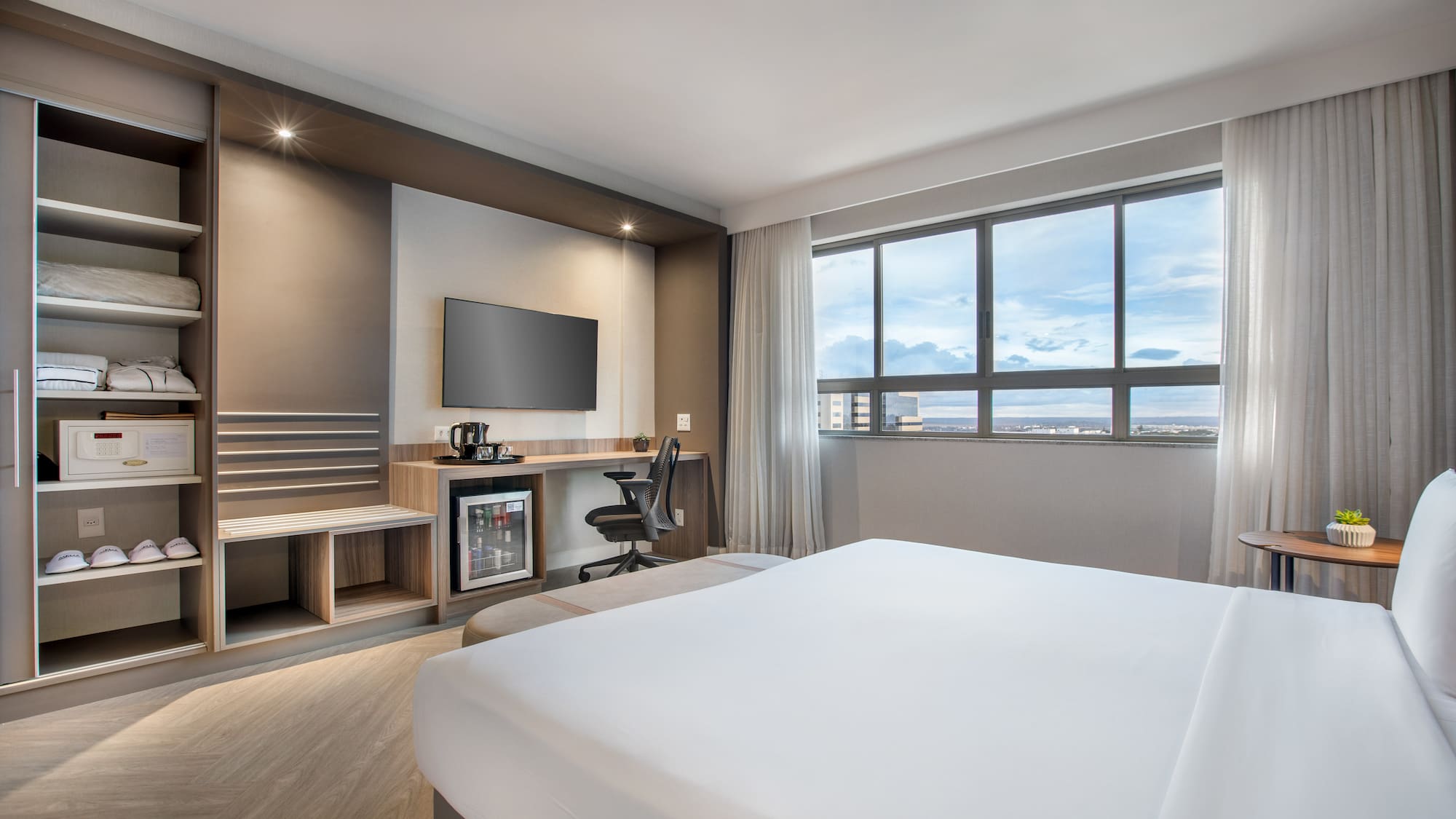 Modern hotel room with bed, workspace, TV, mini-fridge, and city view.