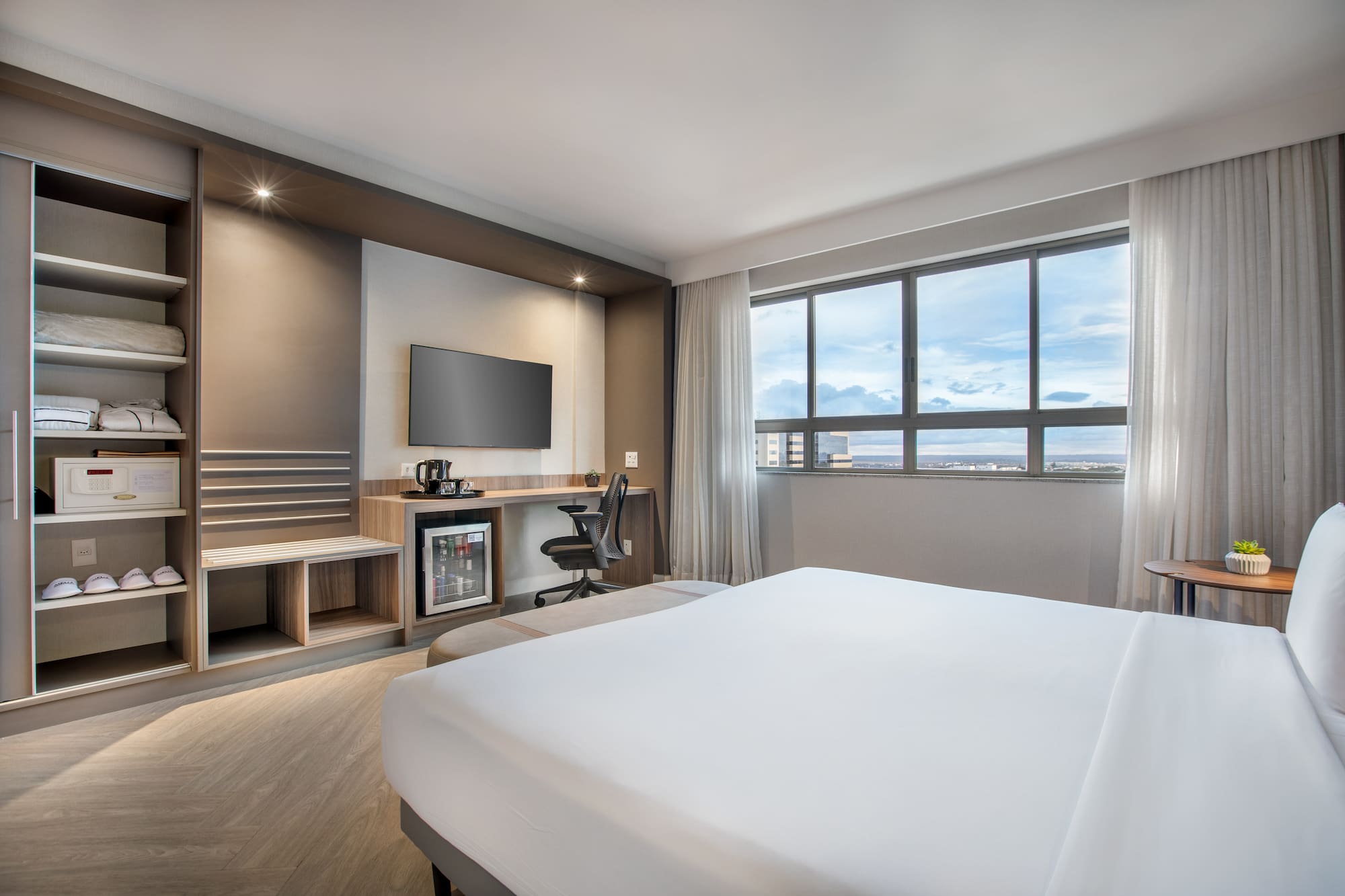 Modern hotel room with bed, workspace, TV, mini-fridge, and city view.