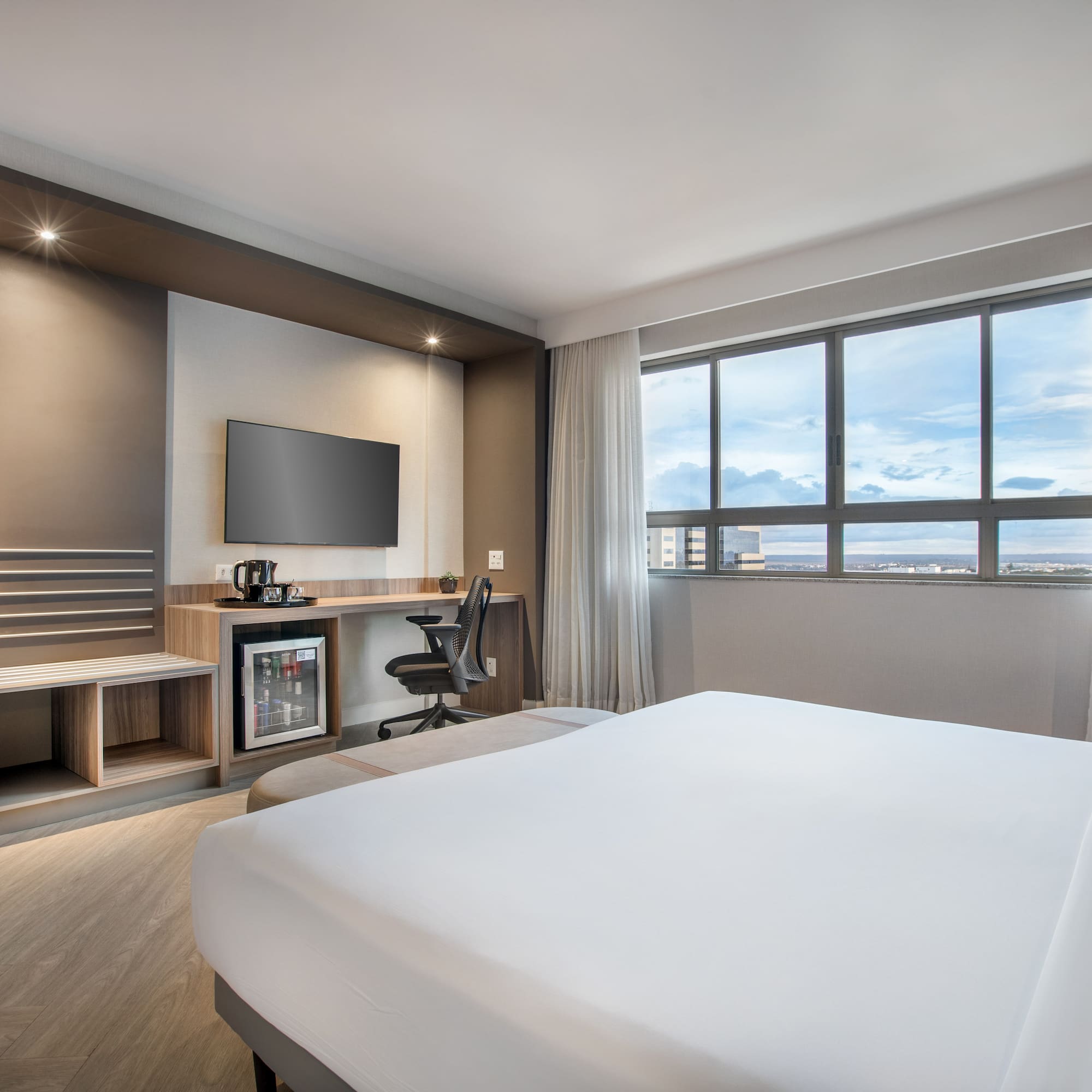 Modern hotel room with bed, workspace, TV, mini-fridge, and city view.