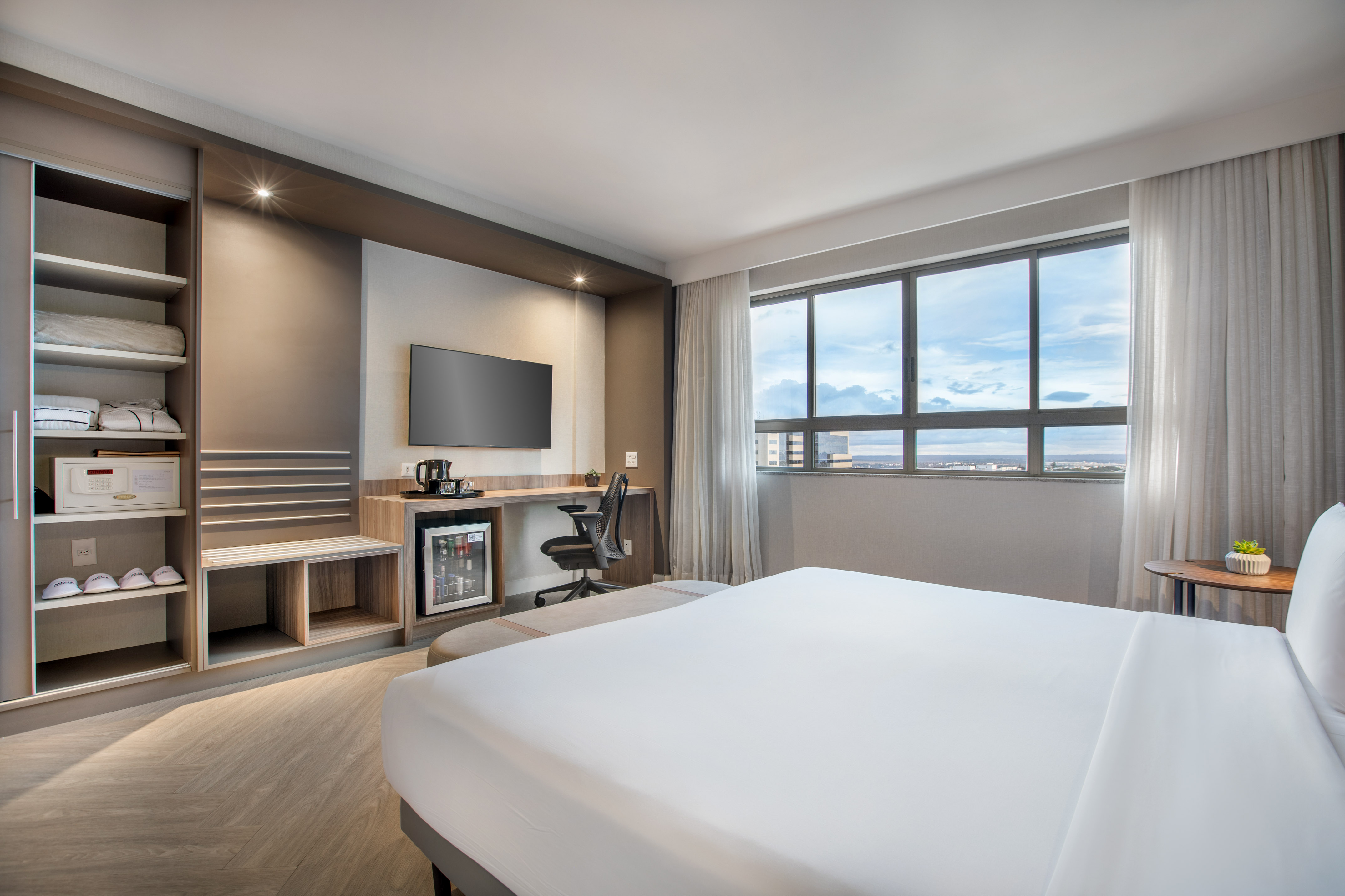 Modern hotel room with bed, workspace, TV, mini-fridge, and city view.