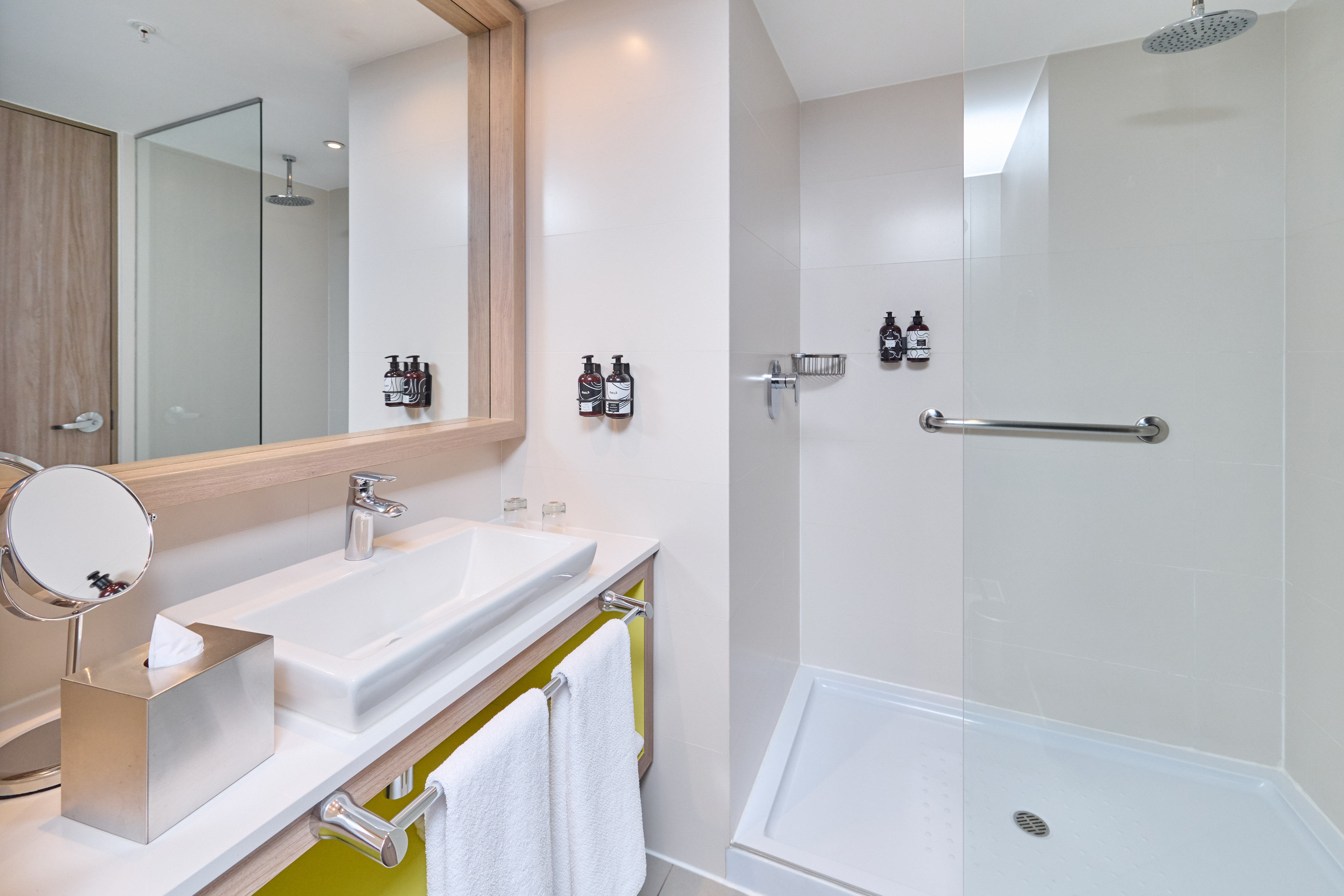 Sleek hotel bathroom: white vanity, mirror, walk-in shower, accessible grab bar.