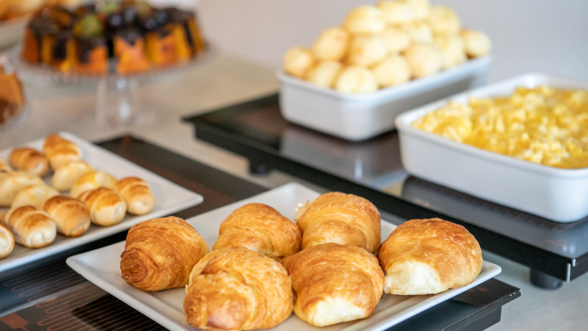 Fresh croissants, pastries, scrambled eggs, and cheese balls on a breakfast buffet.