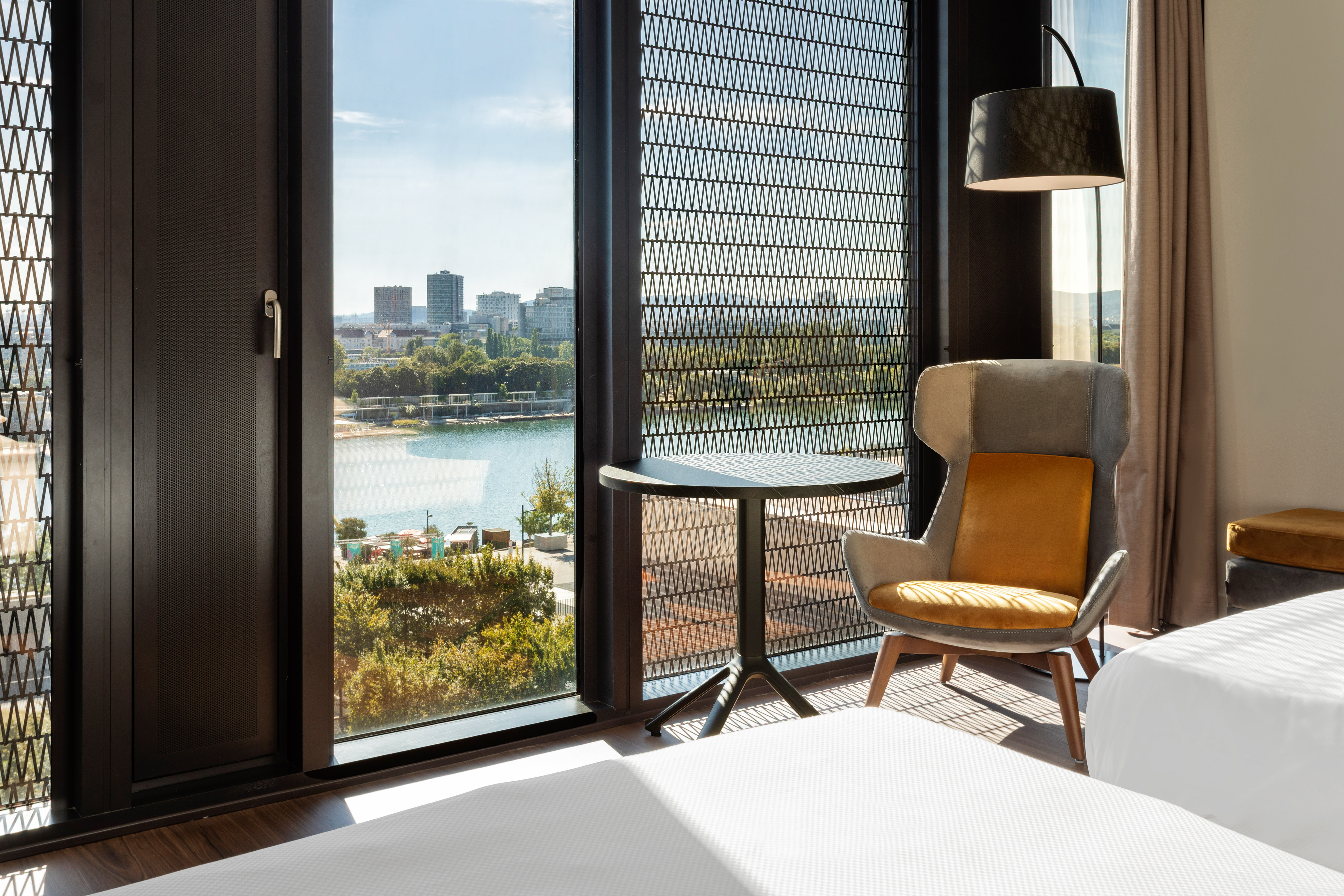 Modern hotel room with city and lake view from large window, featuring armchair.