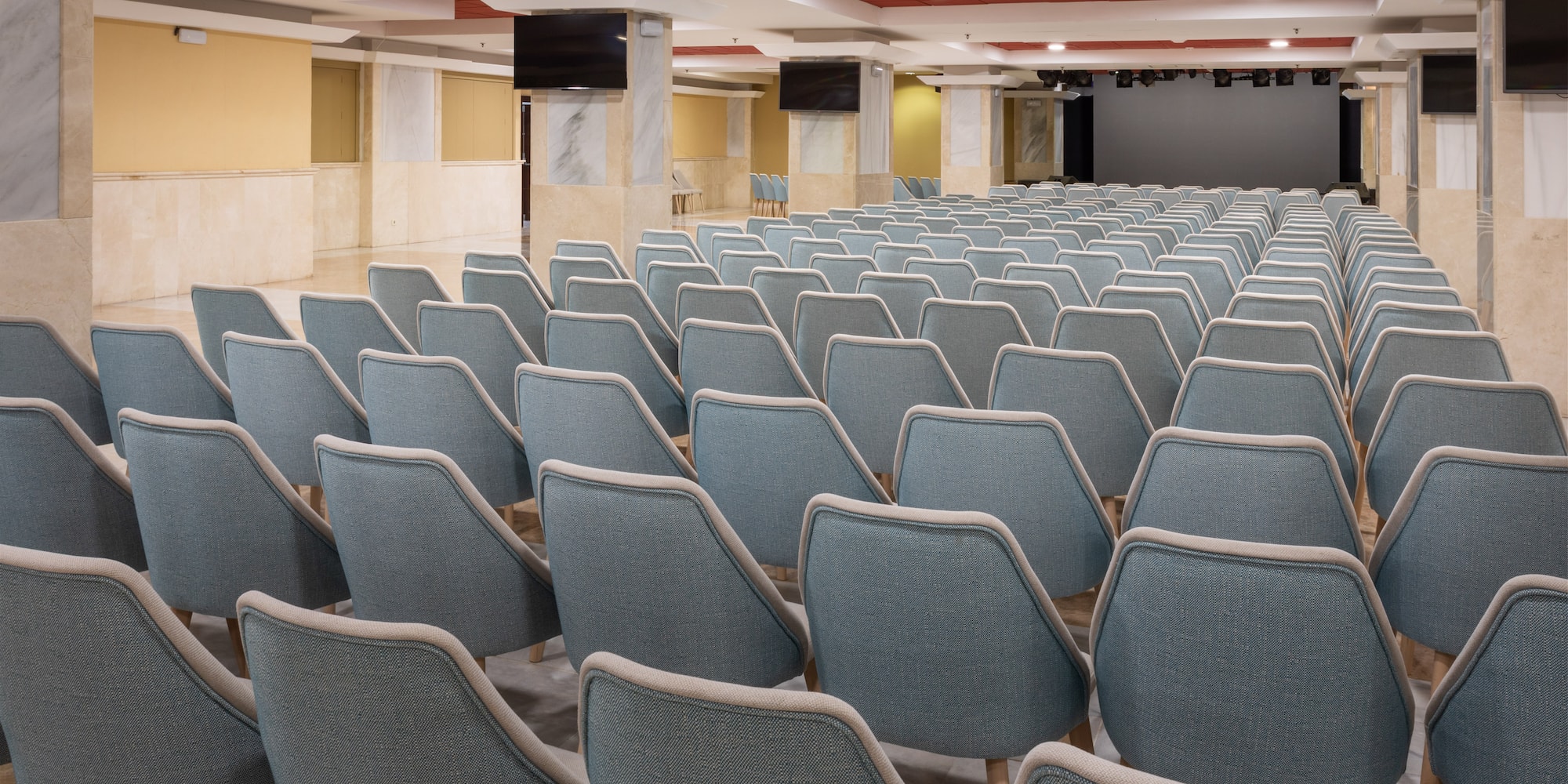 Spacious auditorium with rows of blue chairs facing a stage and screens.