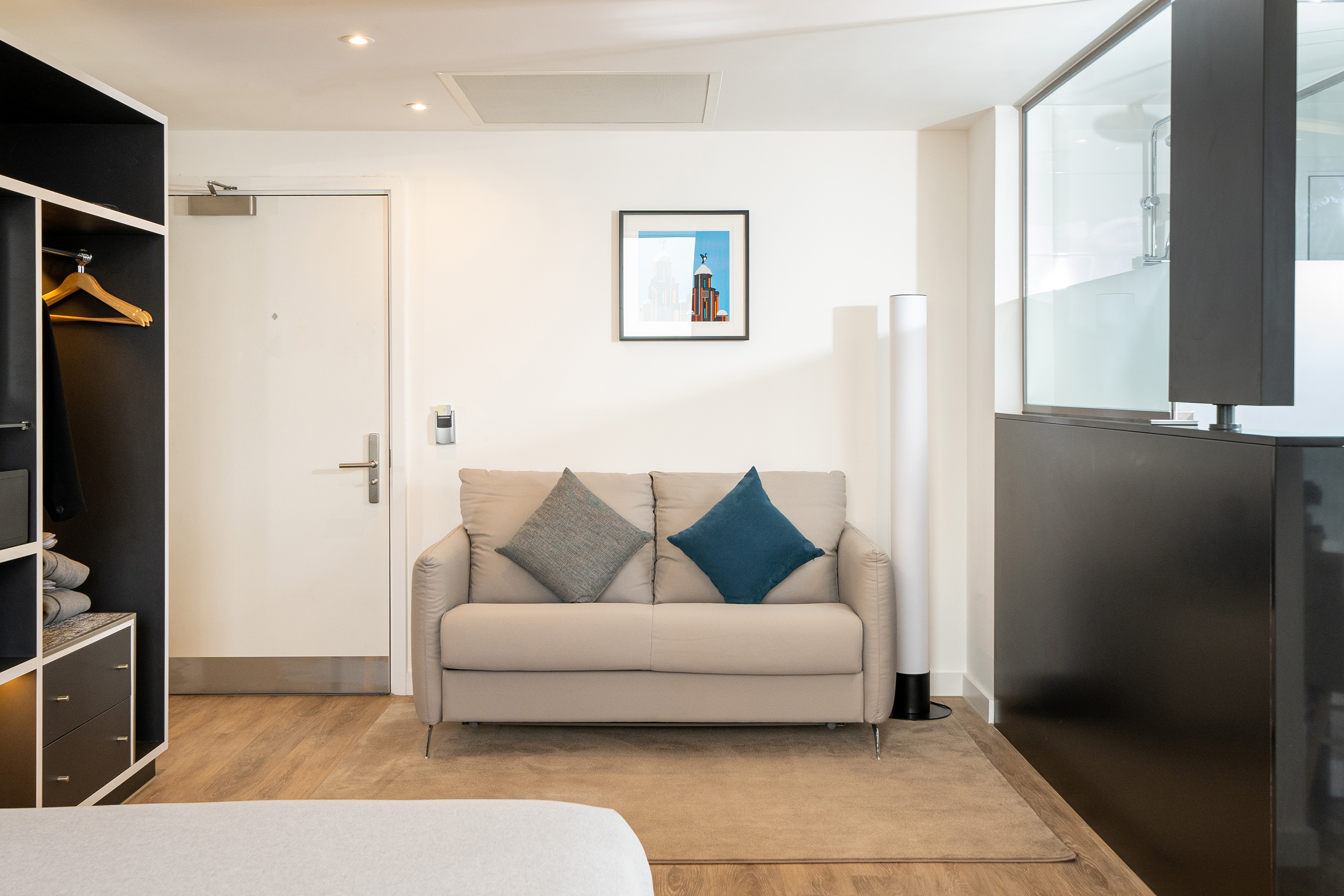 Modern hotel room with beige sofa, open wardrobe, artwork, and glass shower partition.