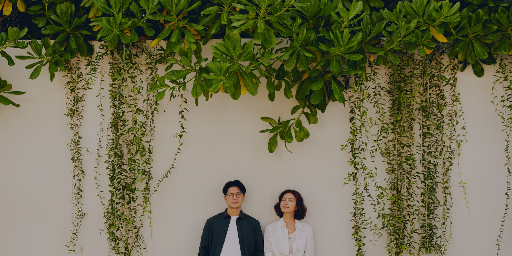 a man and woman standing in front of a wall with plants and trees