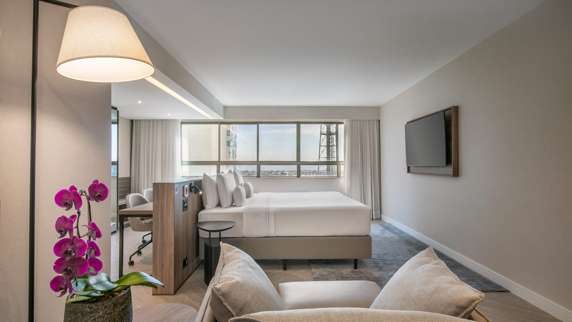 Modern hotel suite with king bed, city view, living area, and desk.
