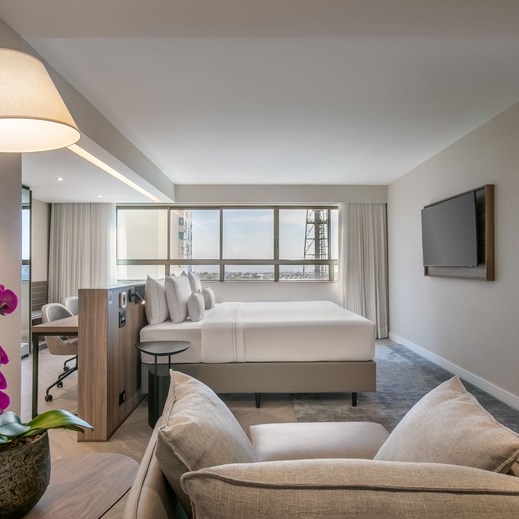 Modern hotel suite with king bed, city view, sofa, and workspace.