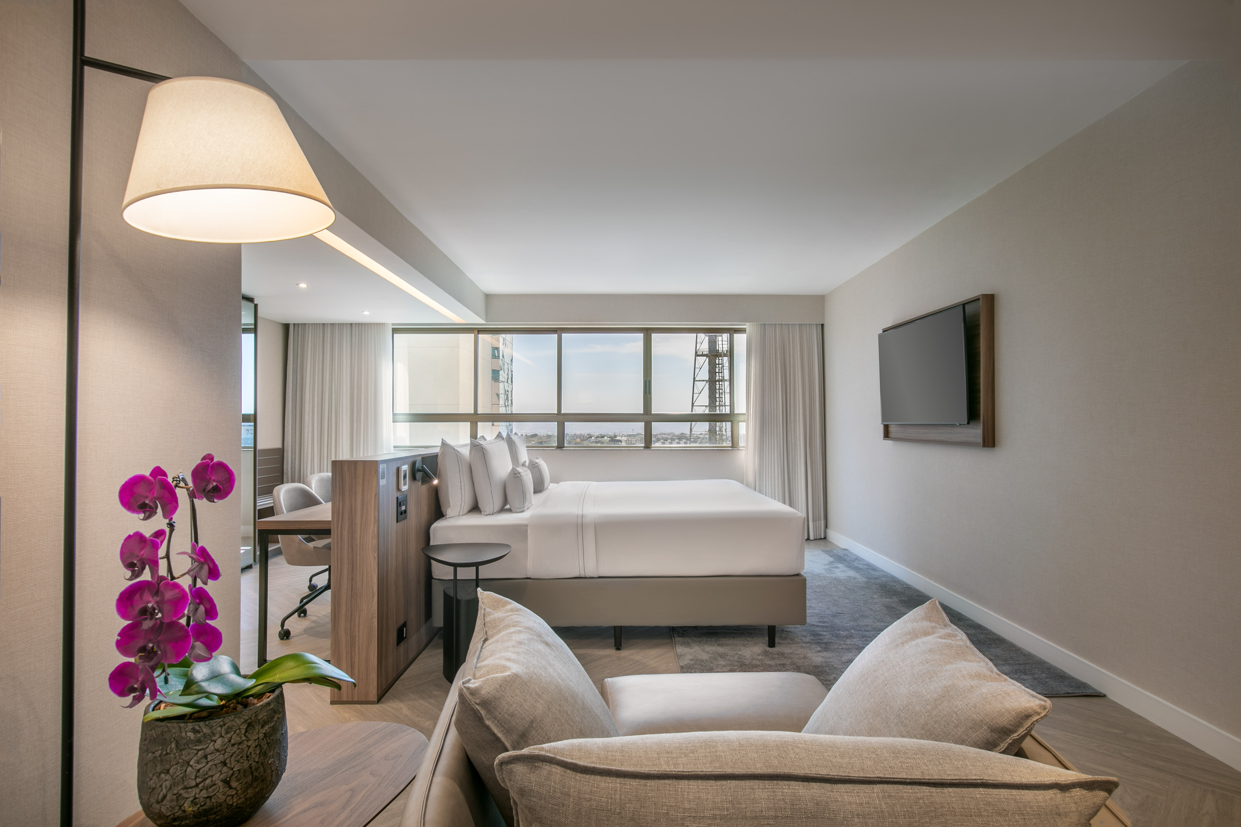 Modern hotel suite with king bed, city view, sofa, and workspace.