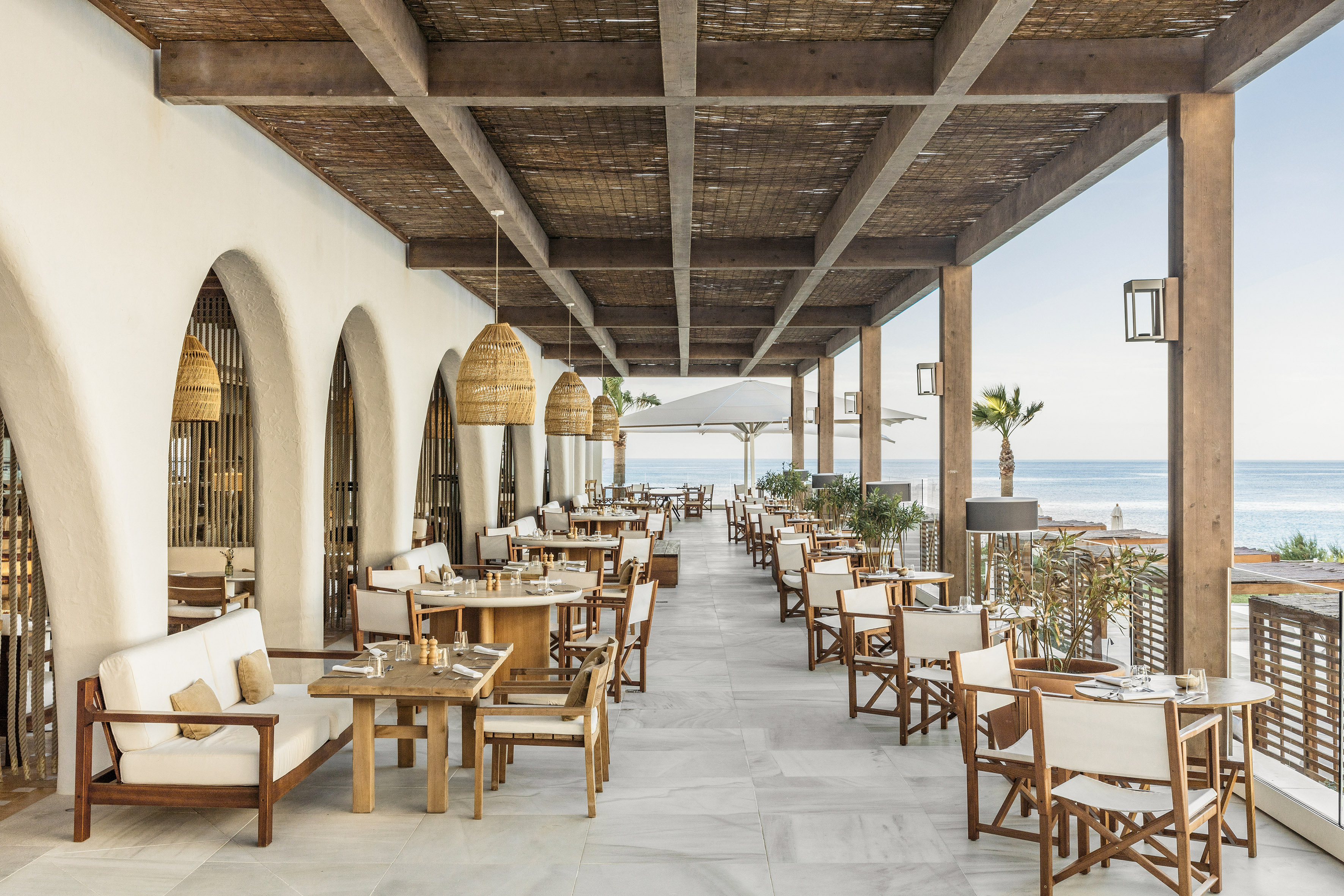 Coastal outdoor restaurant with sea view, wooden tables, and woven ceiling.