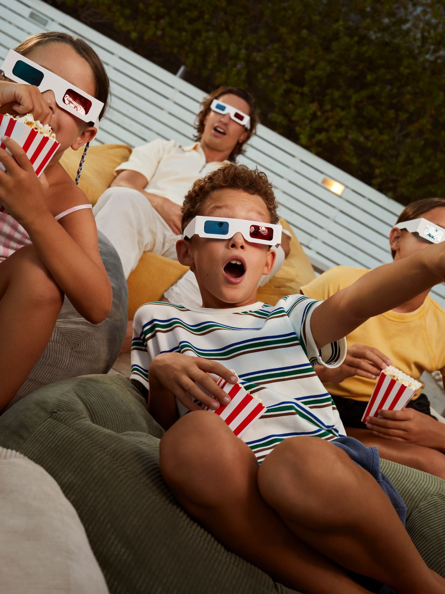 Family group with kids wearing 3D glasses, eating popcorn at outdoor cinema.