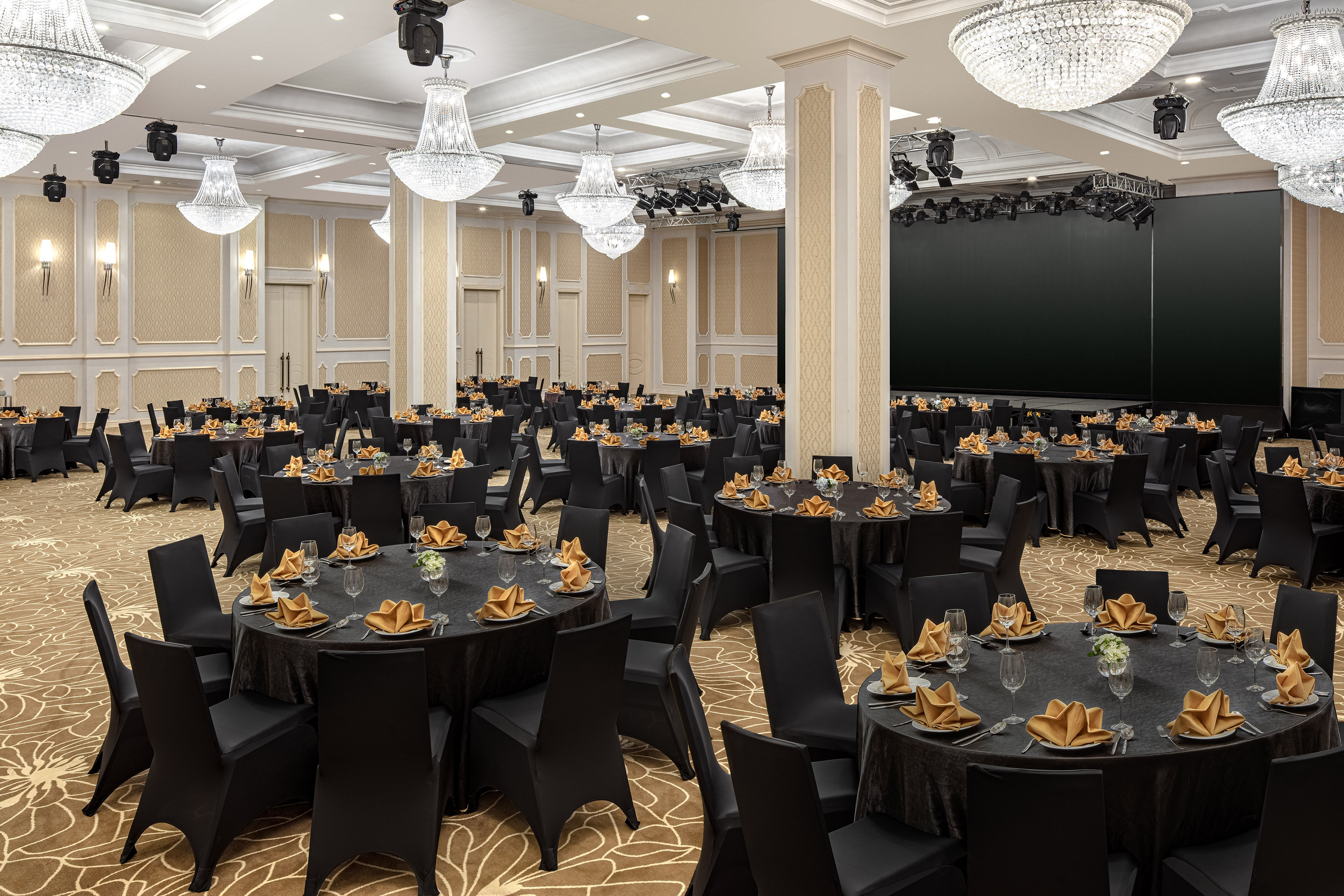 Ballroom set with black tables, gold napkins, crystal chandeliers, large screen.