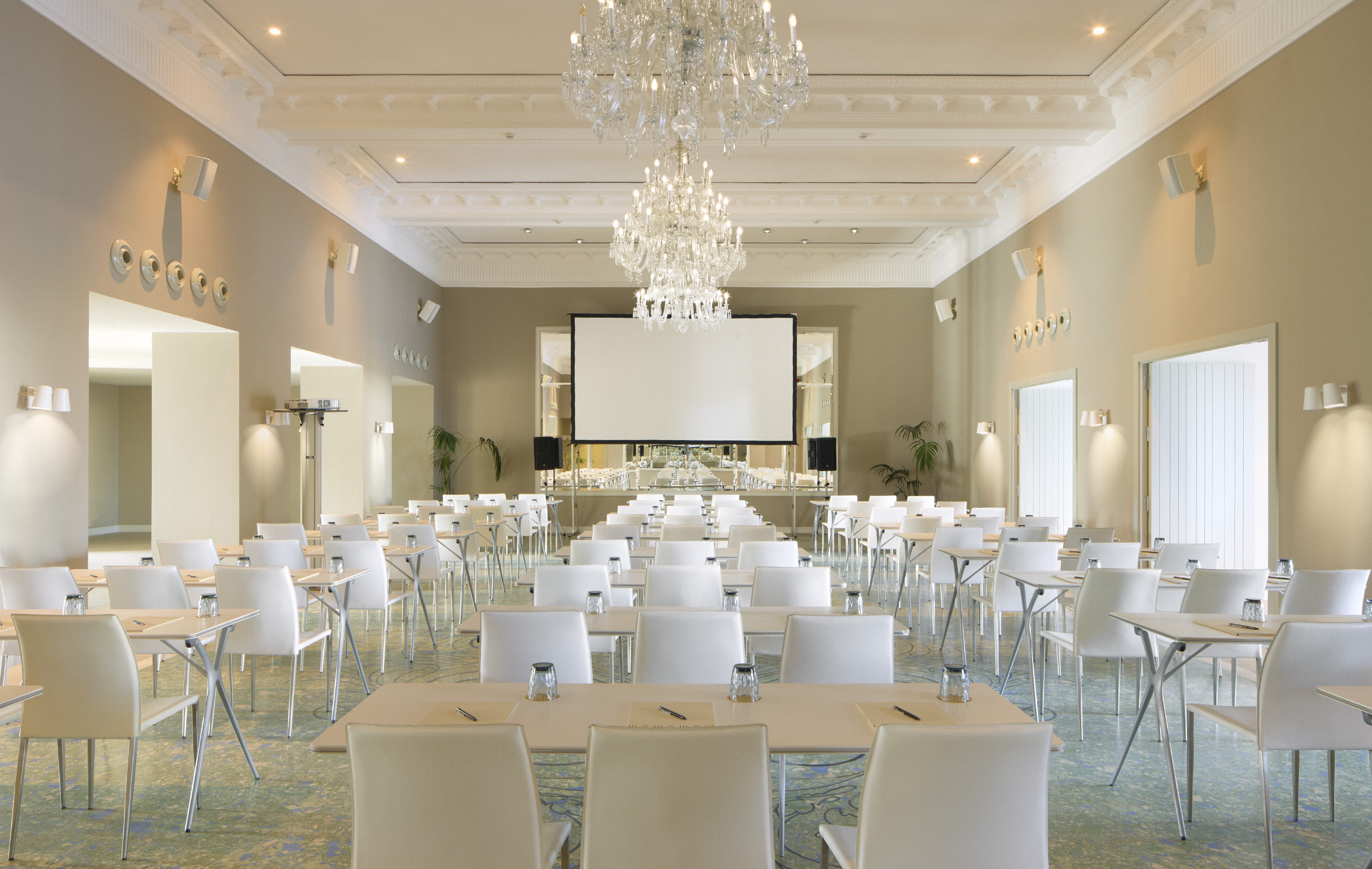 Elegant conference room with projector screen, white tables, and crystal chandeliers.