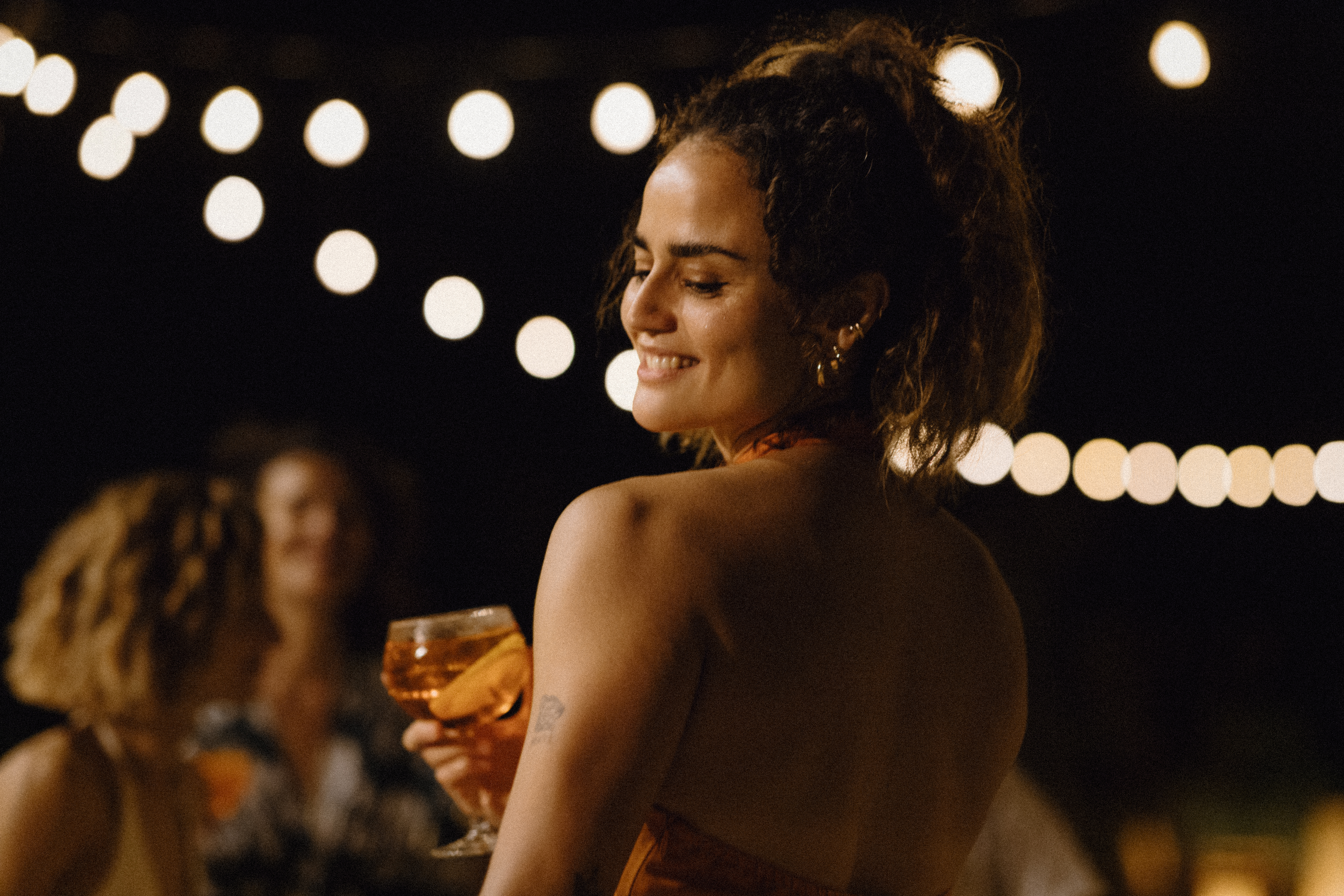 Mujer sonriendo, cóctel en mano, luces festivas de noche.