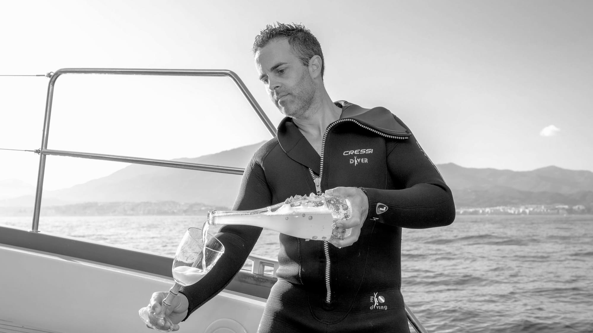 Diver in wetsuit pours champagne on boat, sea and mountains in background.